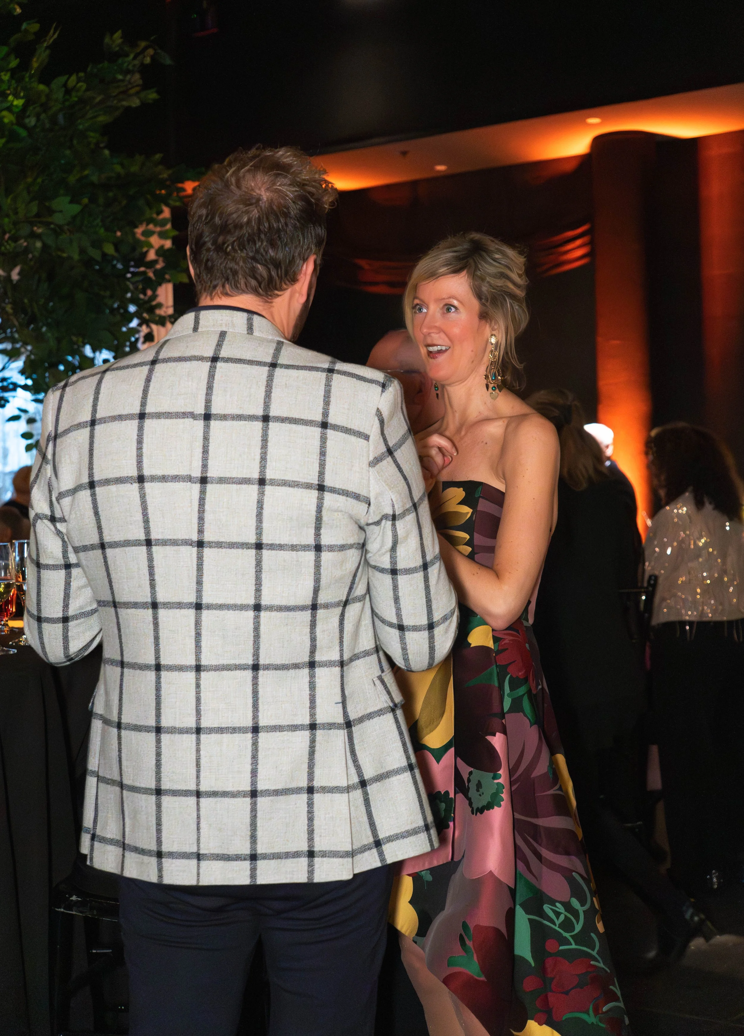 A man and a woman engaging in conversation at a formal event. The woman is wearing a colorful patterned strapless dress and earrings, and appears surprised or excited. The man is dressed in a checked blazer and dark pants. The background has warm lig