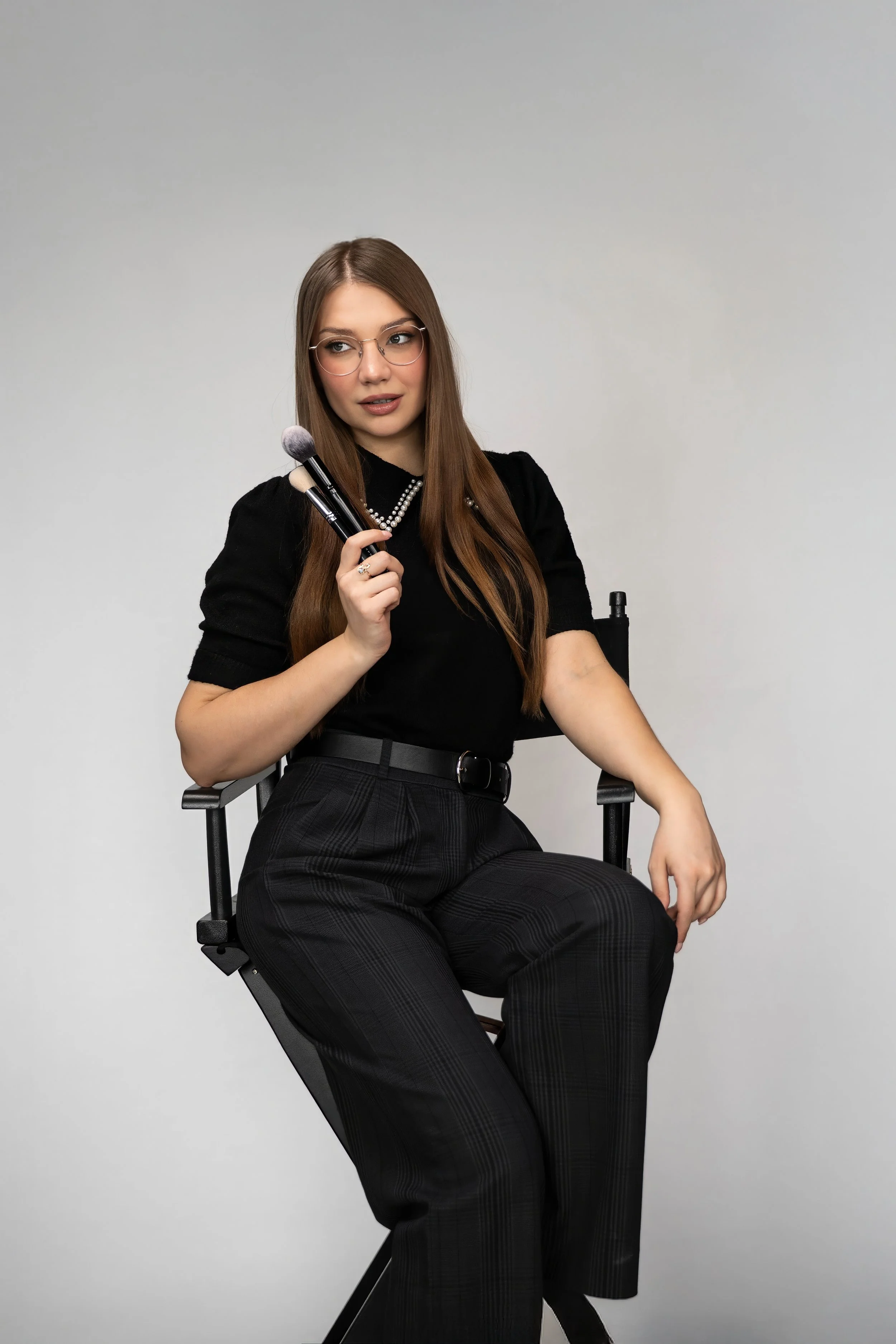 A young woman with long brown hair, wearing glasses, a black shirt with a pearl necklace, and black plaid pants, sitting on a director's chair, holding a makeup brush, against a plain light gray background.