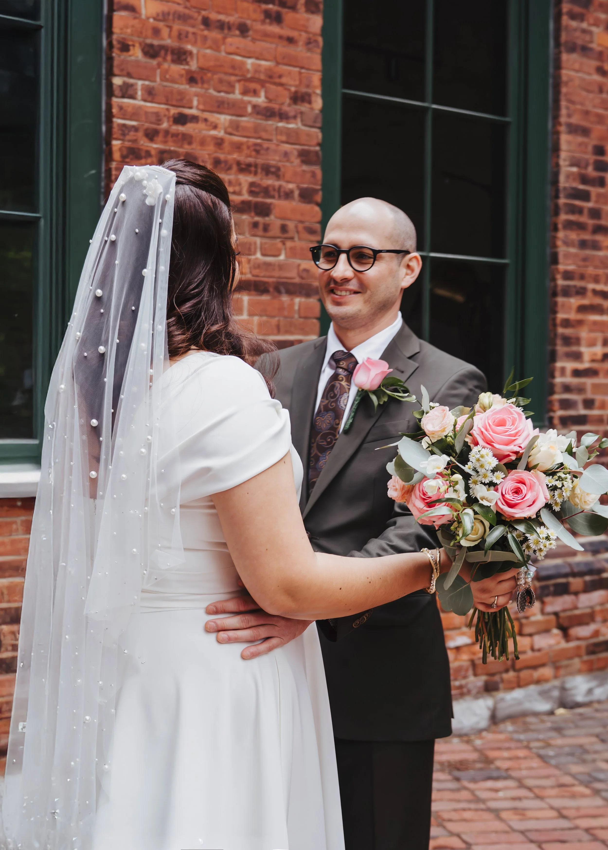 A couple getting married outdoors, with the bride in a white dress and veil holding a bouquet of pink and white roses, and the groom in a suit with a pink rose boutonniere, standing in front of a brick building.