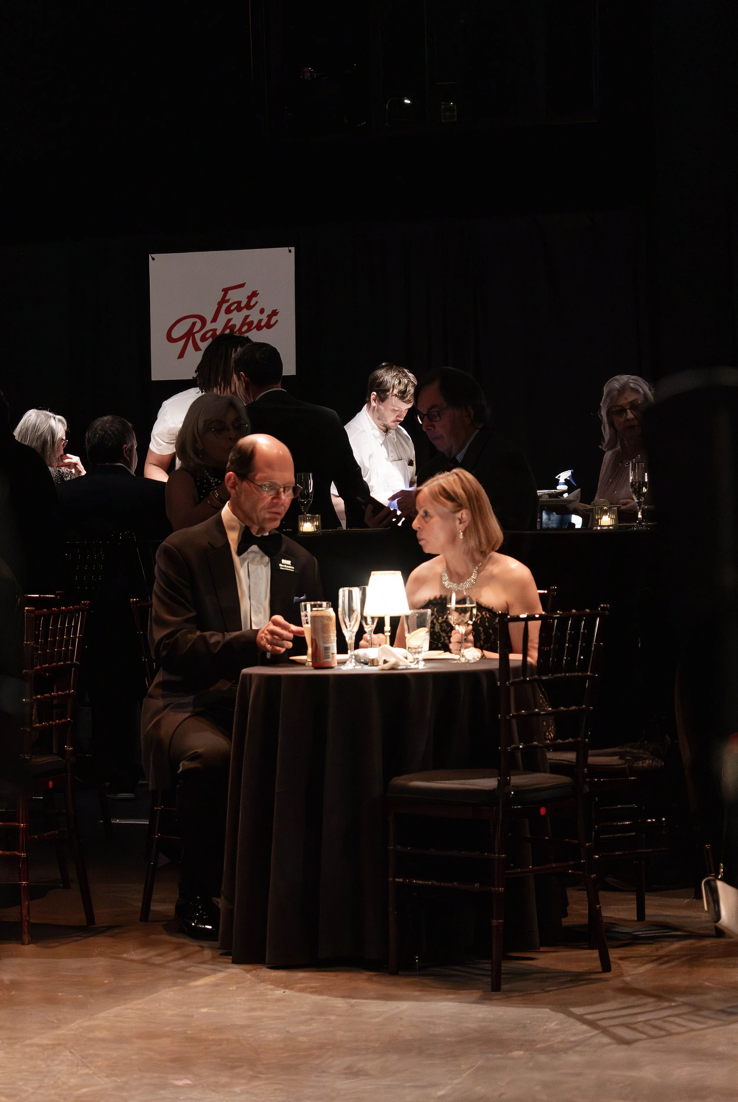 A man and woman in formal attire sitting at a round table with a black tablecloth, having a conversation with drinks and a small lamp, in an elegant event setting with other guests and a wine bar in the background.