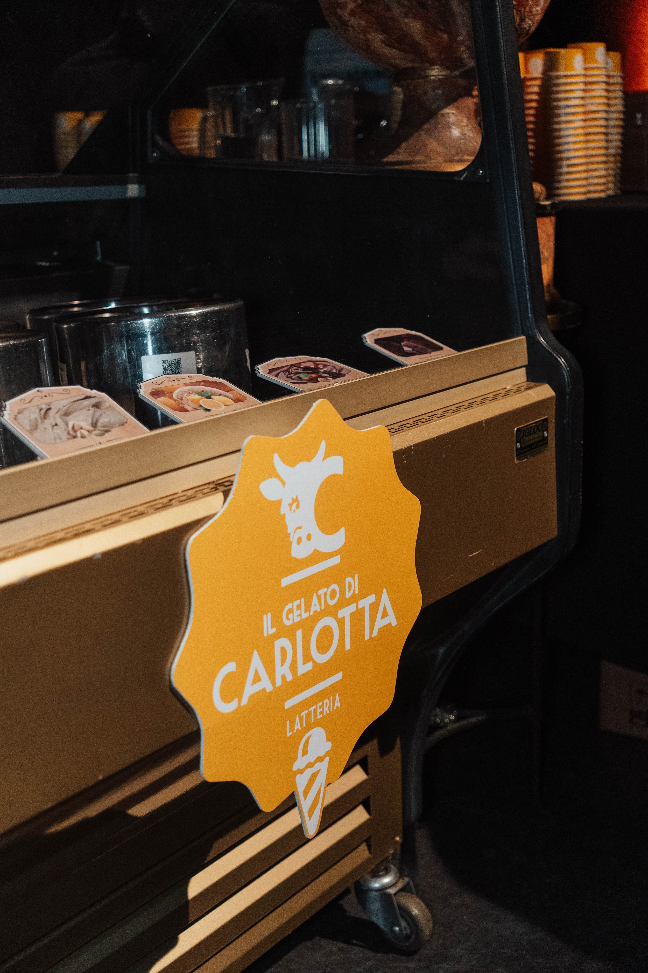 Ice cream freezer with a yellow sign that says "Il Gelato di Carlotta" and features a cow and ice cream cone graphic. Inside the freezer, ice cream flavors are visible in small containers, with some printed labels on top.