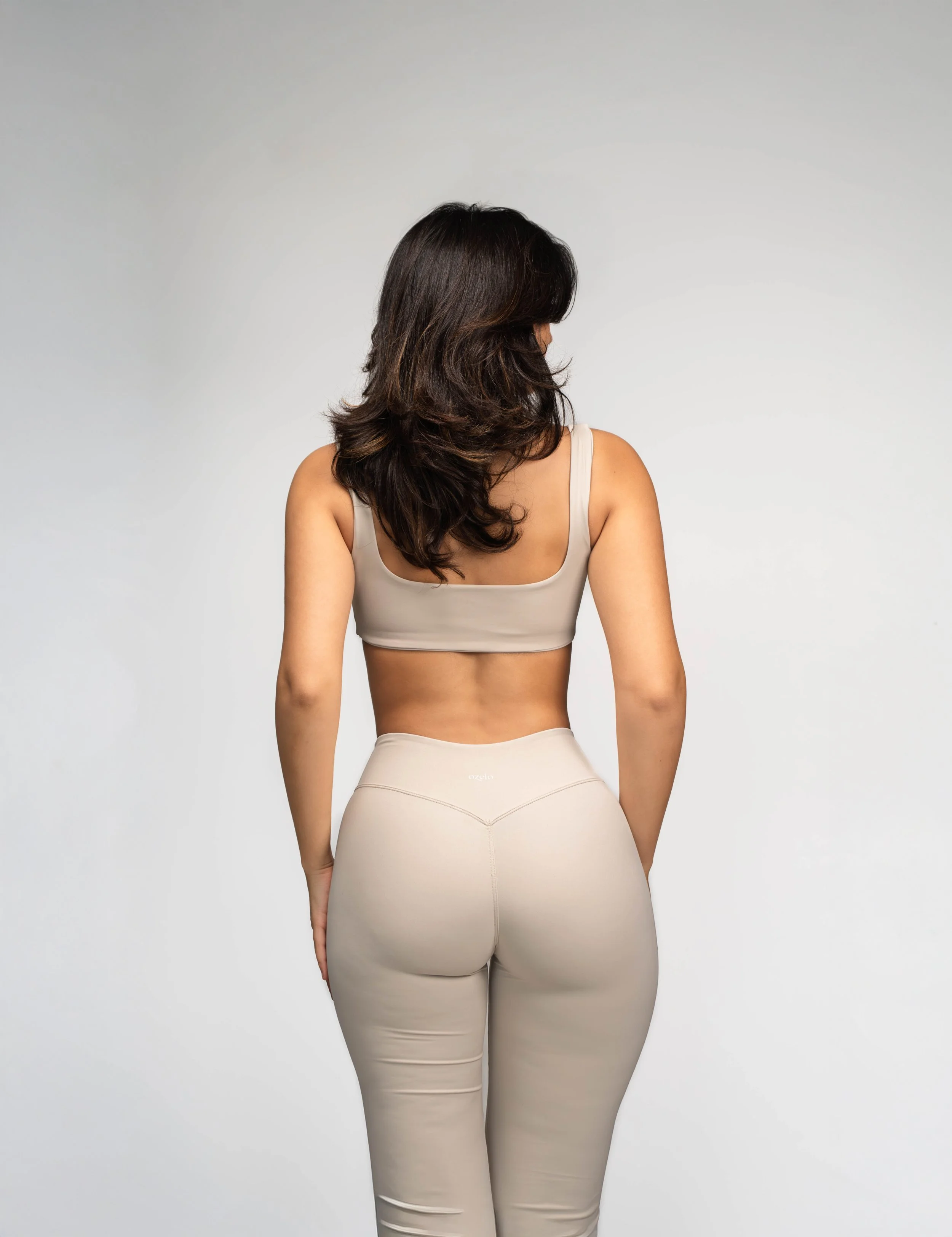 A woman with dark, wavy hair wearing a beige sports bra and matching high-waisted leggings, facing away against a plain gray background.
