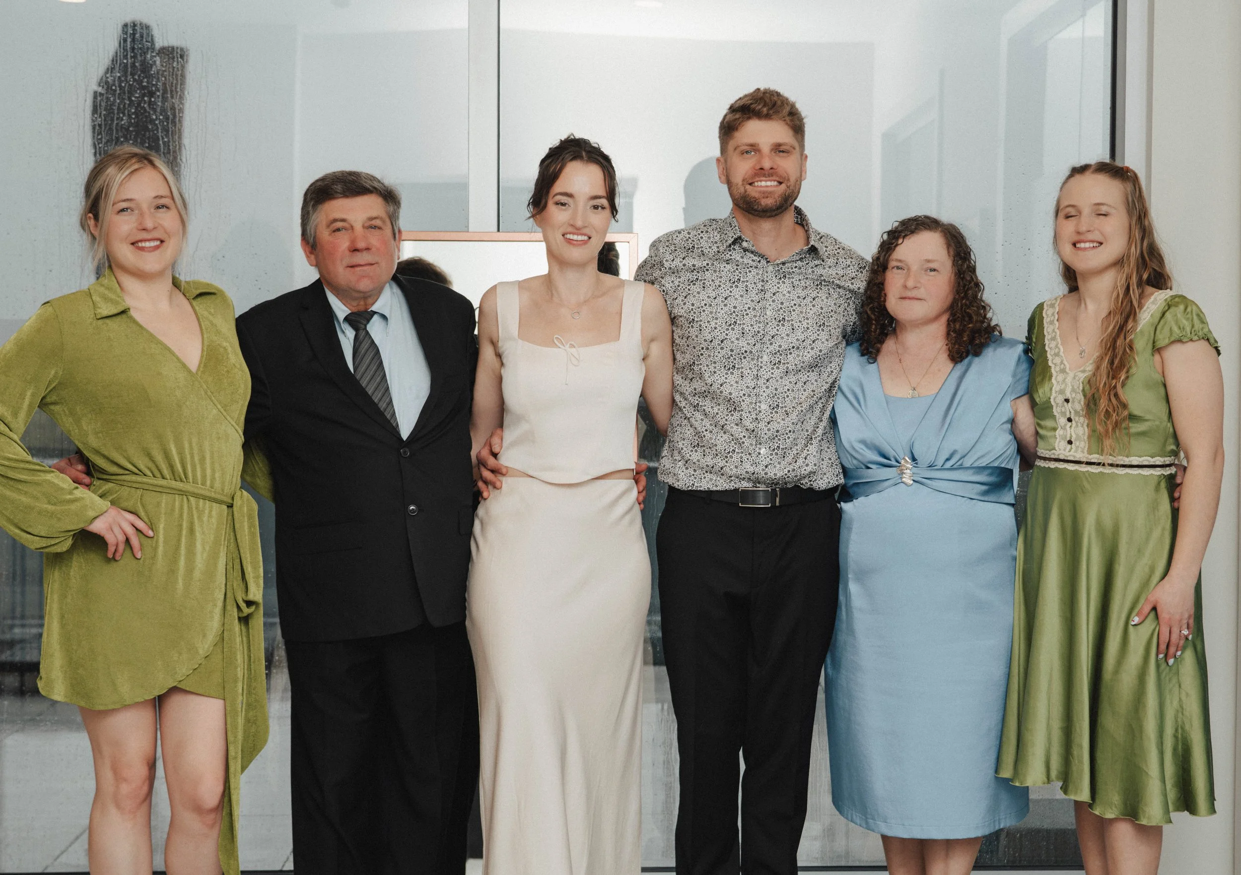 Group of seven people standing in front of a glass wall, smiling and posing for a photo. The group consists of three women and four men of various ages, dressed in casual and semi-formal clothing.