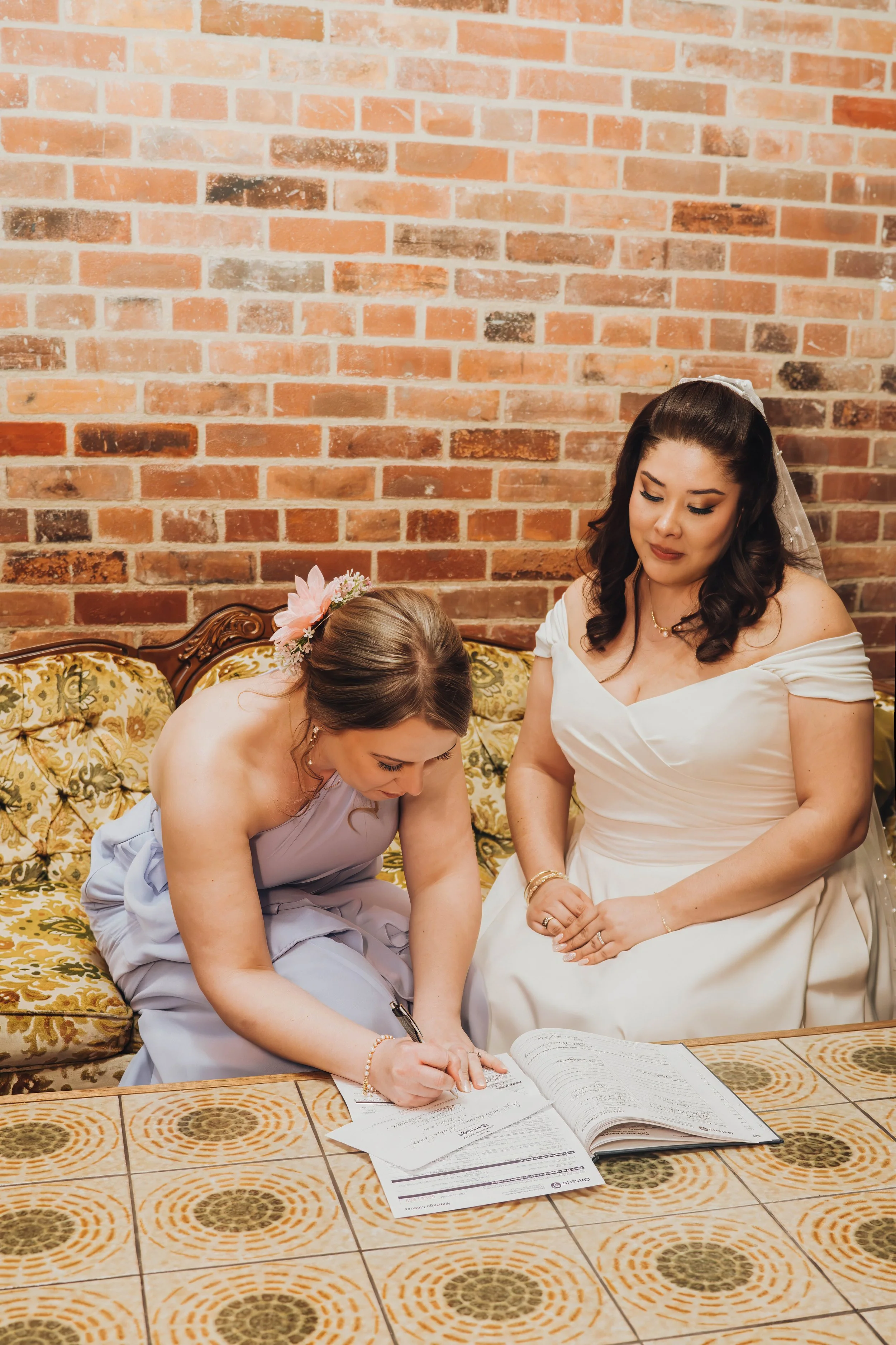 A woman in a wedding dress is seated on a vintage sofa, while another woman in a lavender dress is signing a document on a tiled table. The background features a brick wall.