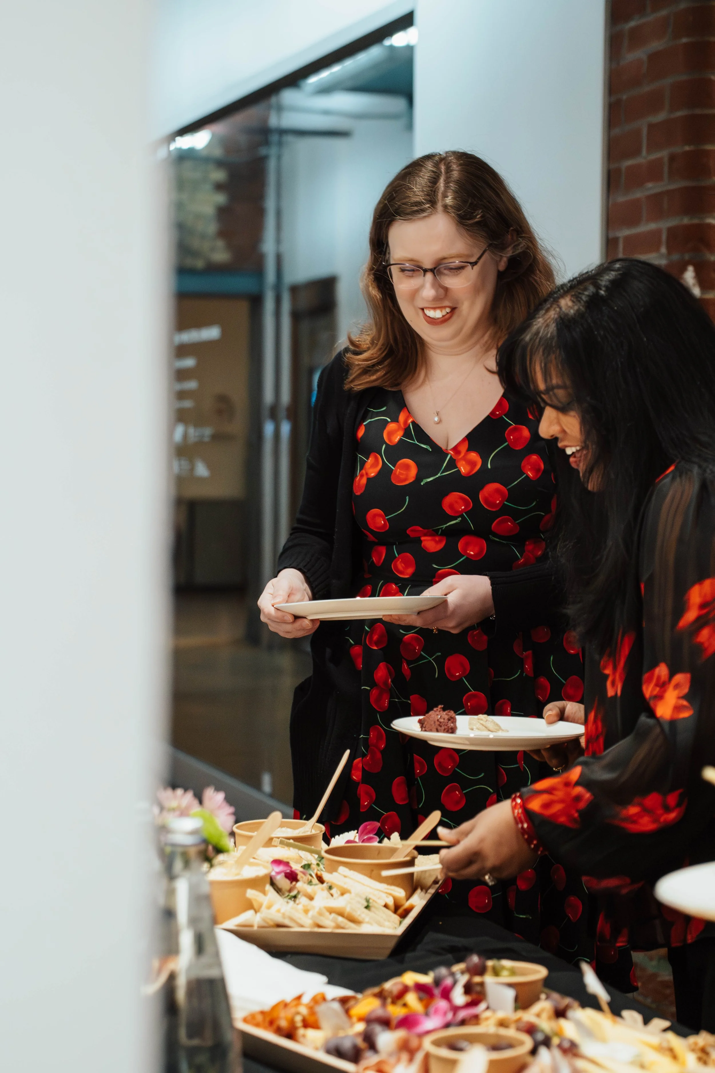Two women smiling and serving themselves at a buffet table with various foods, wearing matching cherry-patterned dresses.