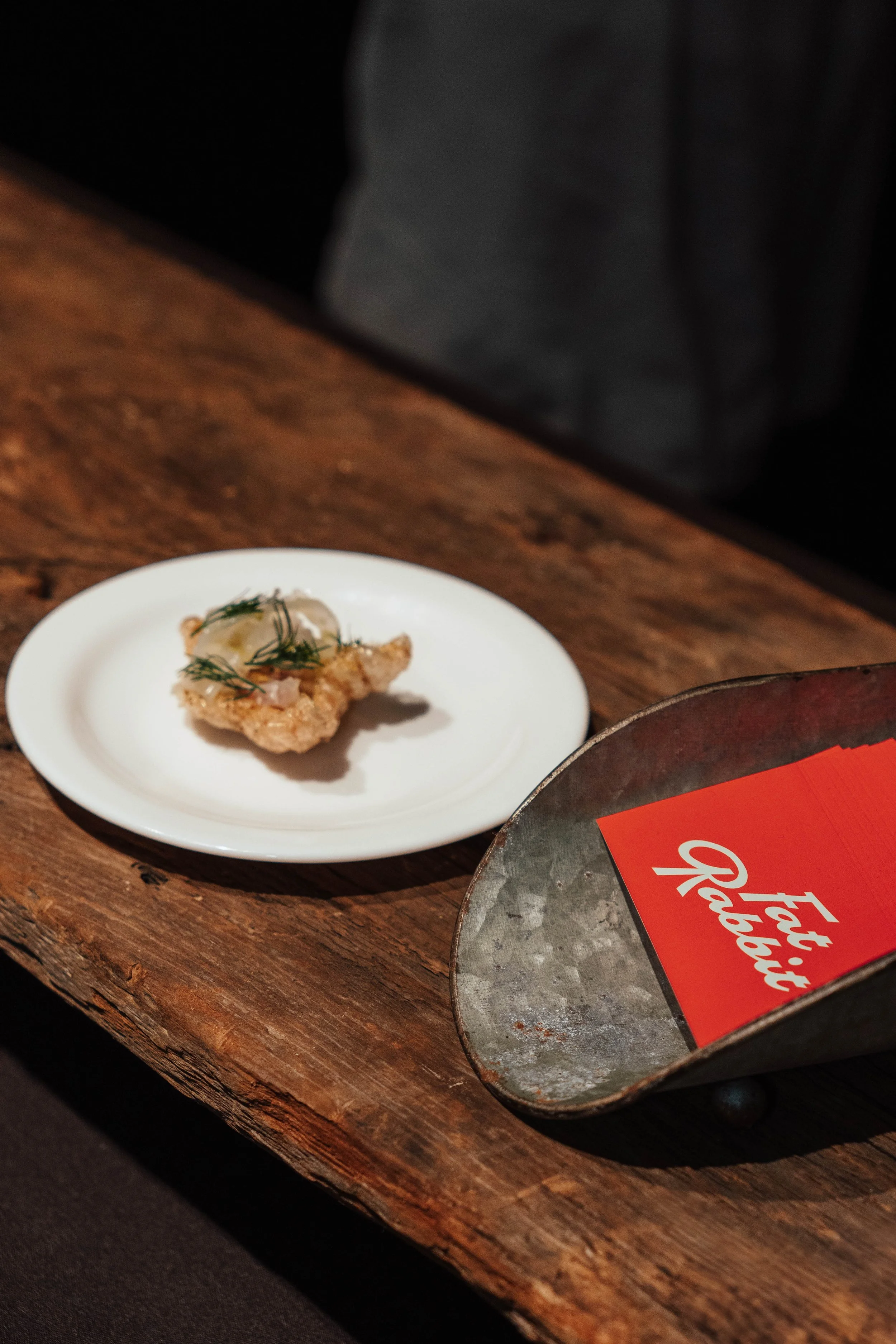 A small piece of fried food garnished with herbs on a white plate, placed on a rustic wooden table, next to a metal tray holding a red packet with white text.