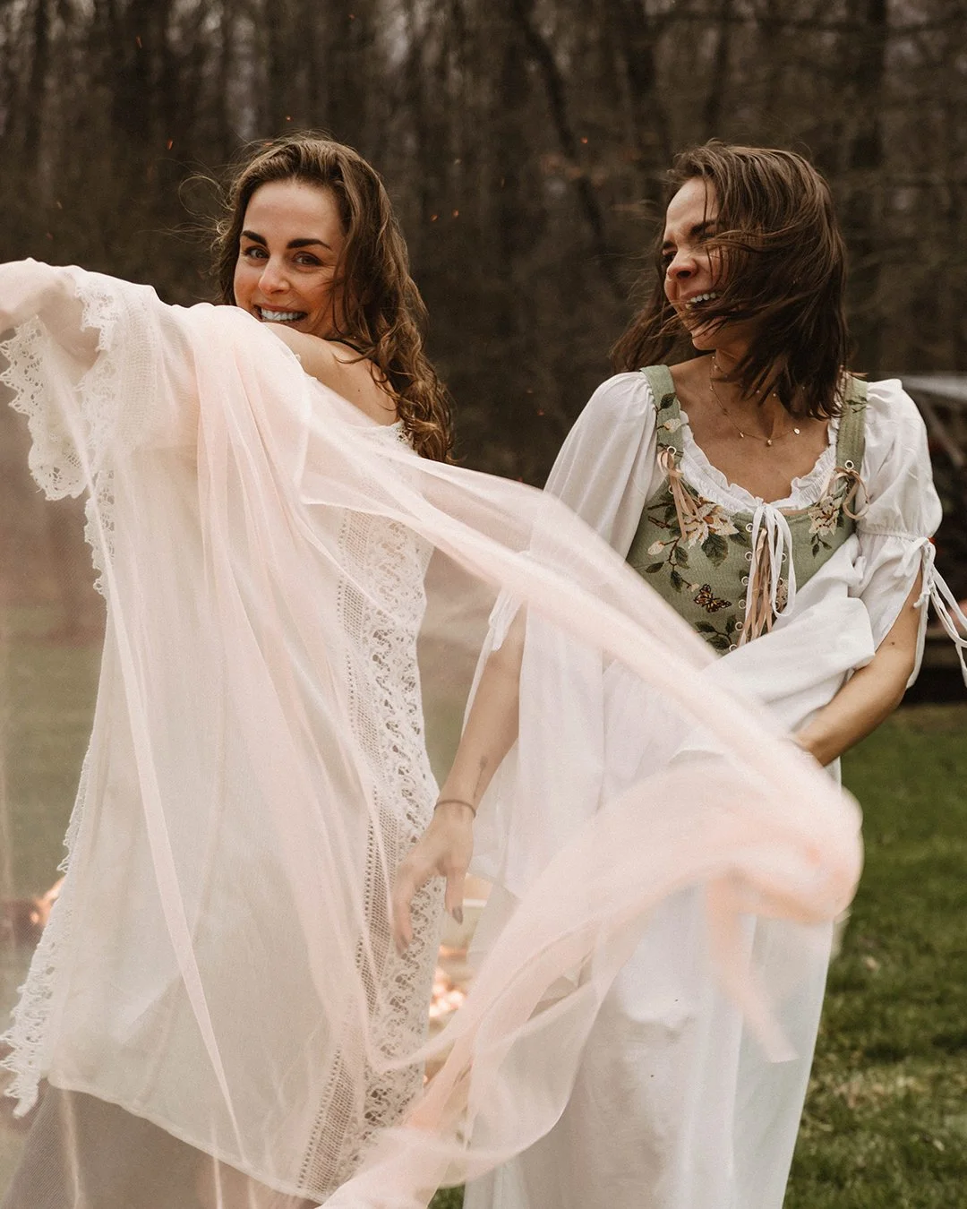 Two women dancing outdoors during sunset, one in a white lace dress and the other in a floral dress with puffed sleeves, both smiling and enjoying themselves.