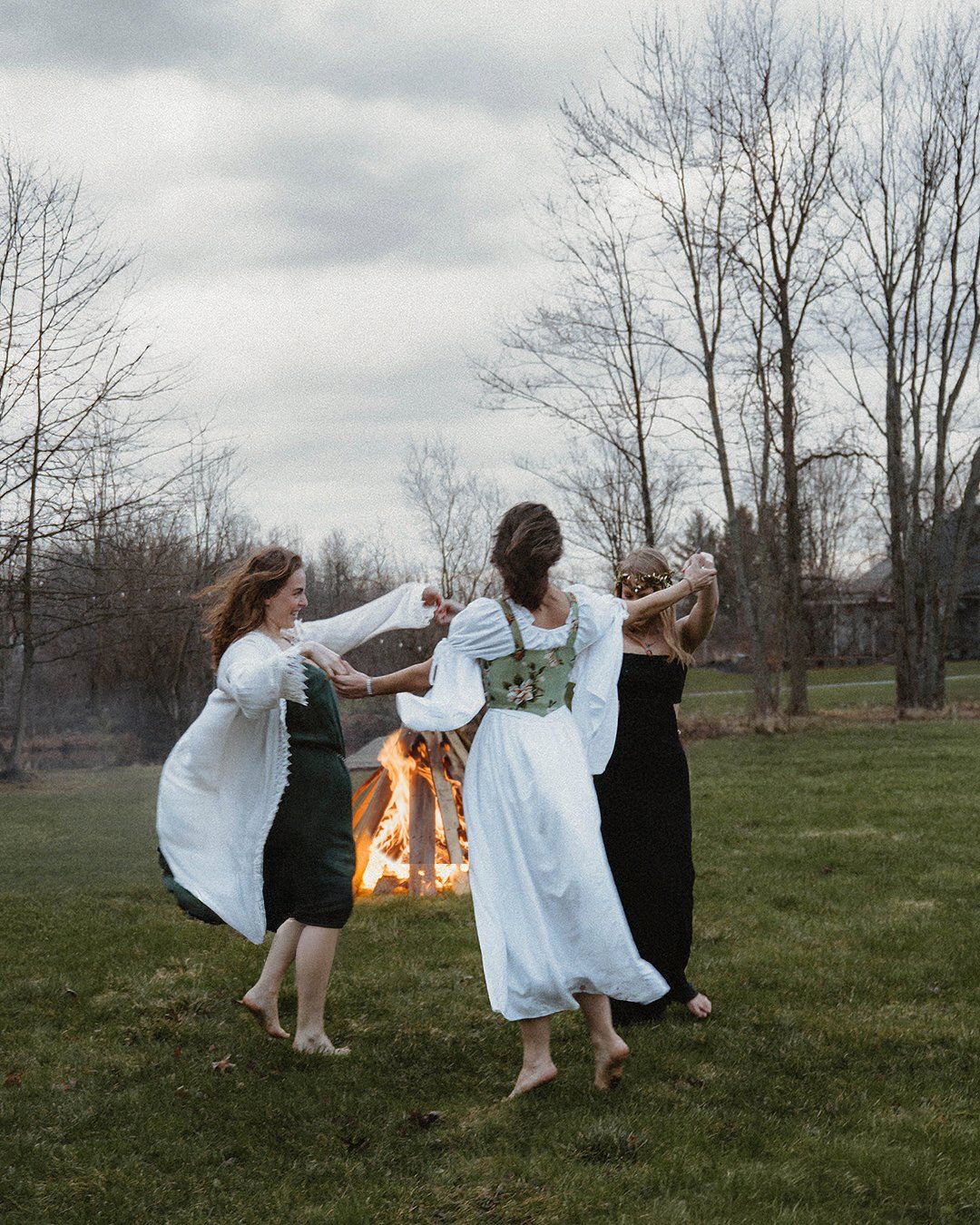 Four women in bohemian clothing dancing in a circle around a bonfire outdoors during overcast weather, with leafless trees in the background.