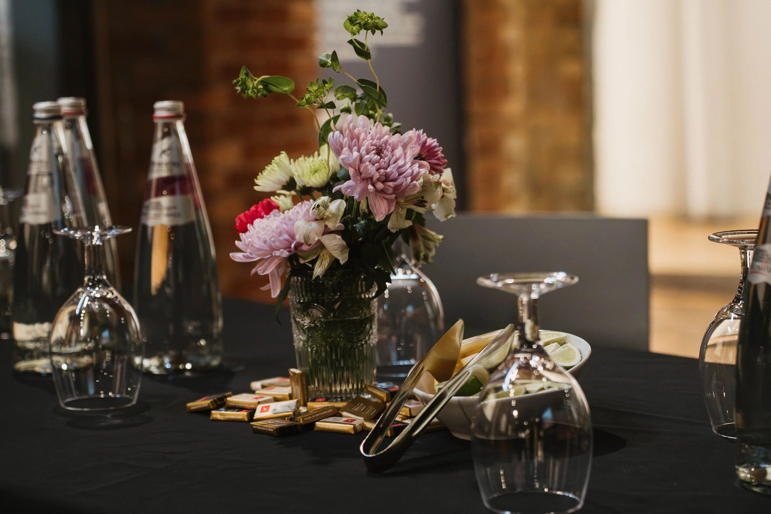 A table with a black tablecloth, decorated with a floral arrangement of pink, white, and red flowers in a glass vase, surrounded by upside-down wine glasses, a bowl of lime wedges, and small packages of butter or chocolates.
