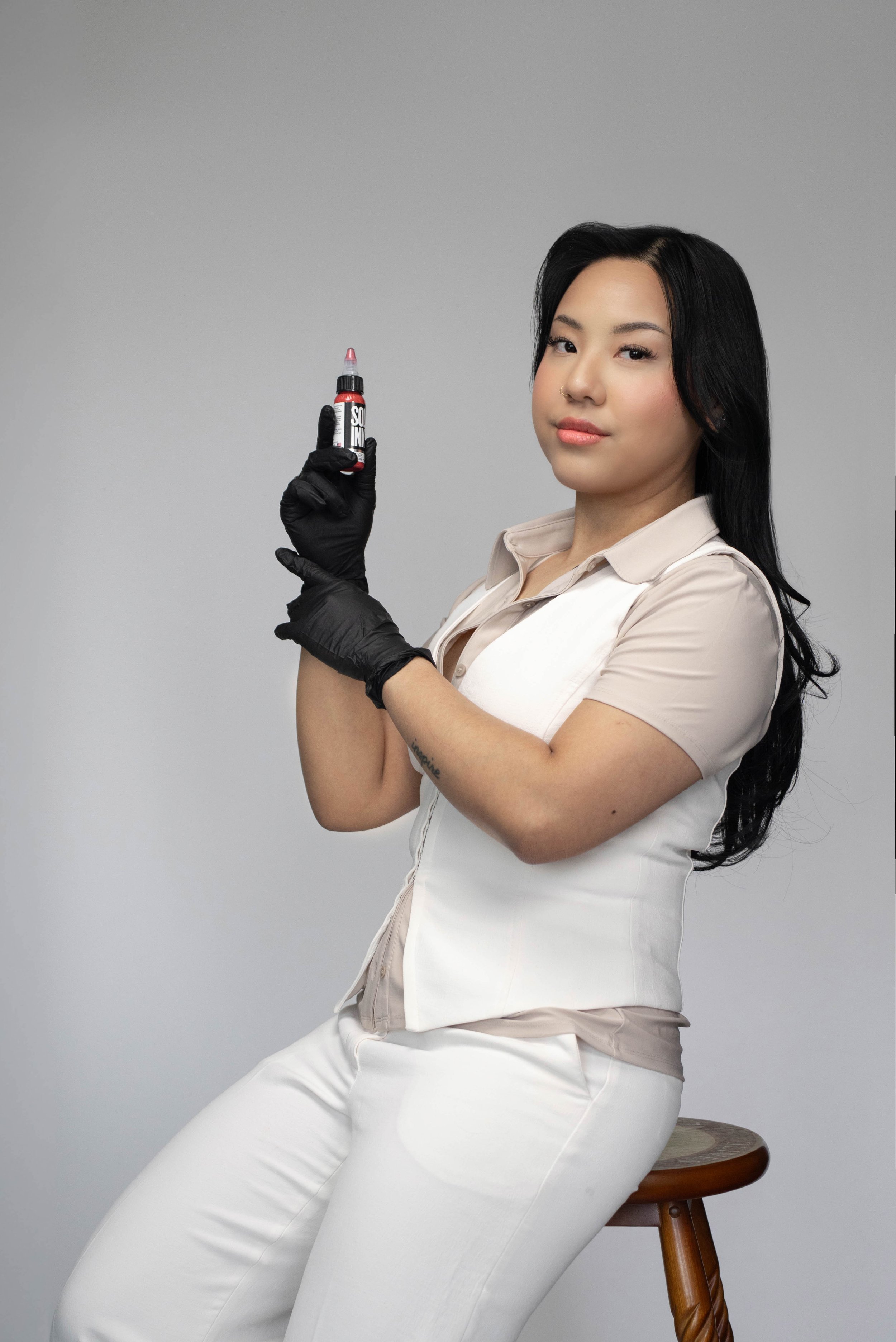 A woman with long black hair, wearing a beige short sleeve shirt and white pants, sits on a wooden stool against a plain gray background. She is holding a small bottle with a red and black label and a pink tip, wearing black gloves, and slightly smil