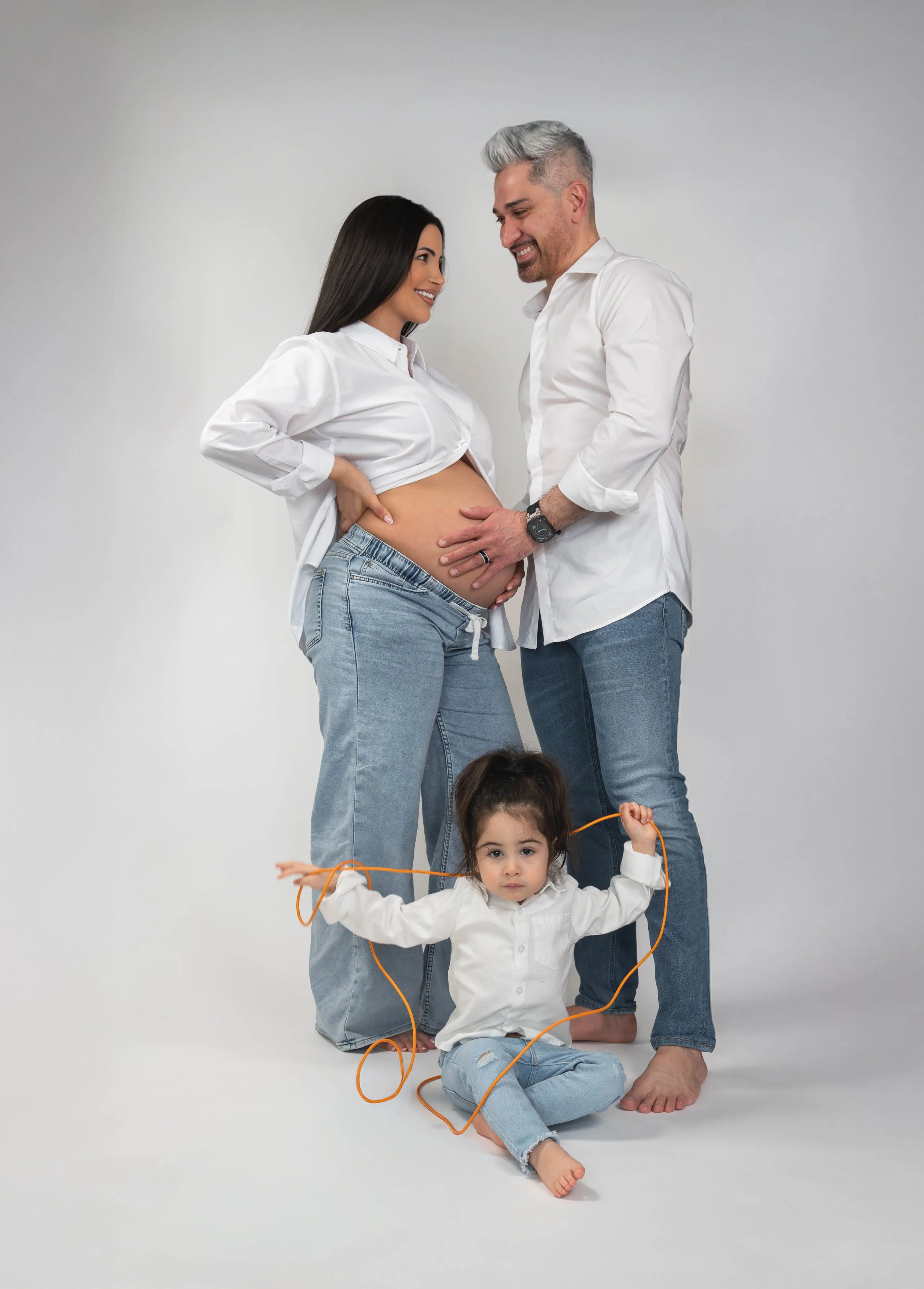 A pregnant woman and a man are standing close together, smiling, with the woman's belly exposed, as she is wearing unbuttoned white shirt and jeans. A young girl is sitting on the floor in front of them, holding an orange jump rope, with a neutral ex