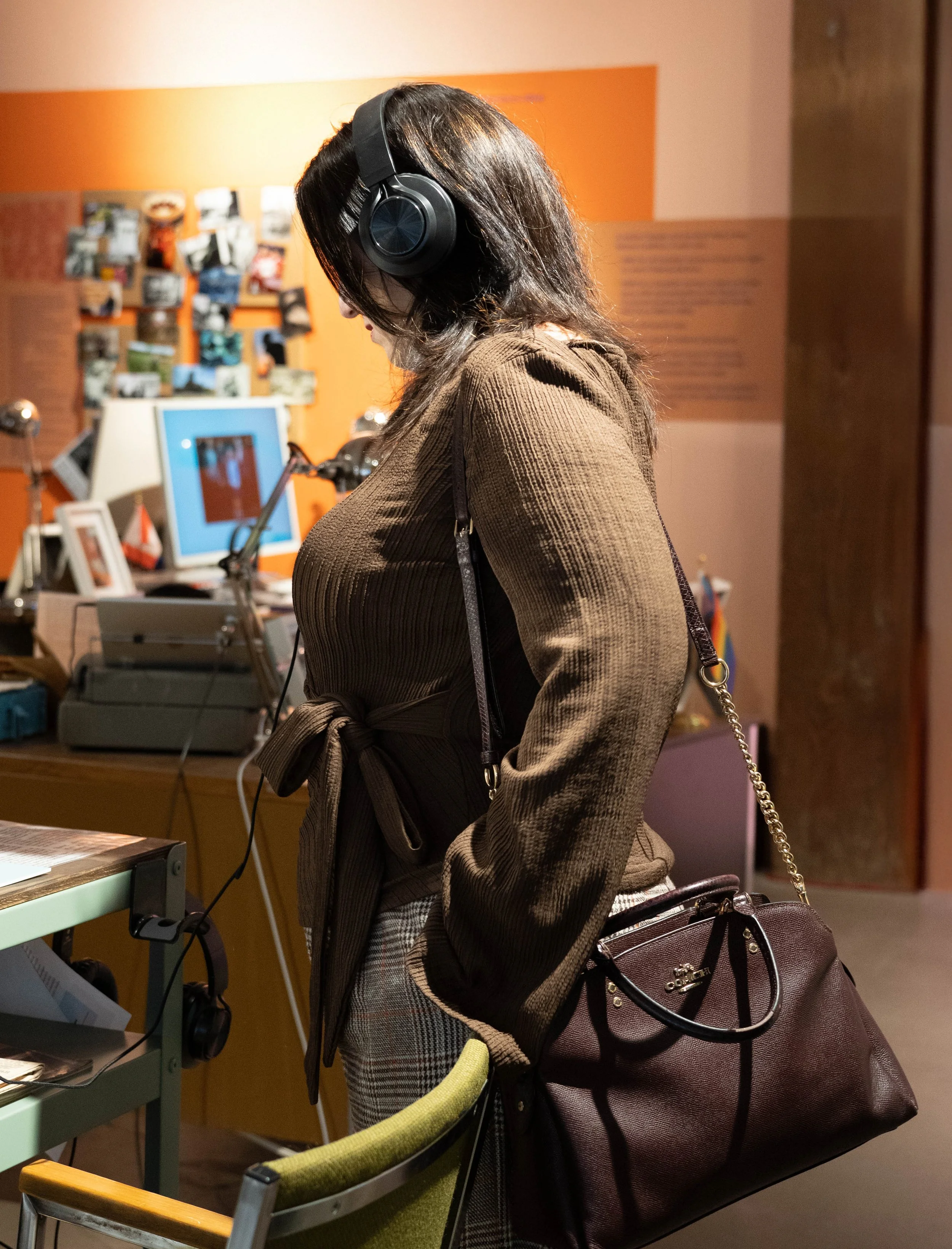 A woman with brown hair wearing large black headphones, a brown belted coat, and carrying a brown purse, standing in a room with orange and pink walls, surrounded by computers, photographs, and office supplies.