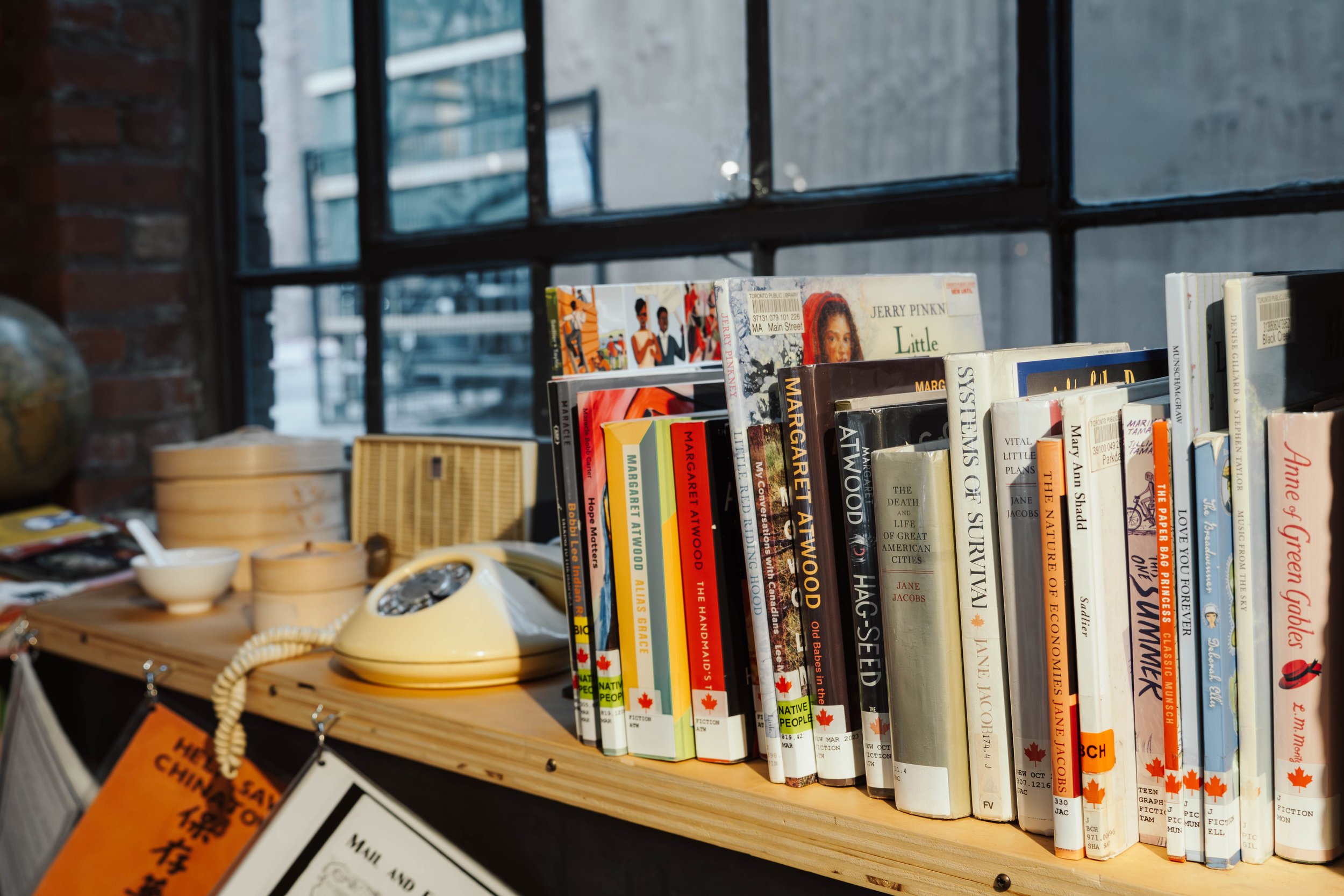 A wooden table with a vintage telephone, a bowl, and book on it, set against a large window with a brick wall outside.