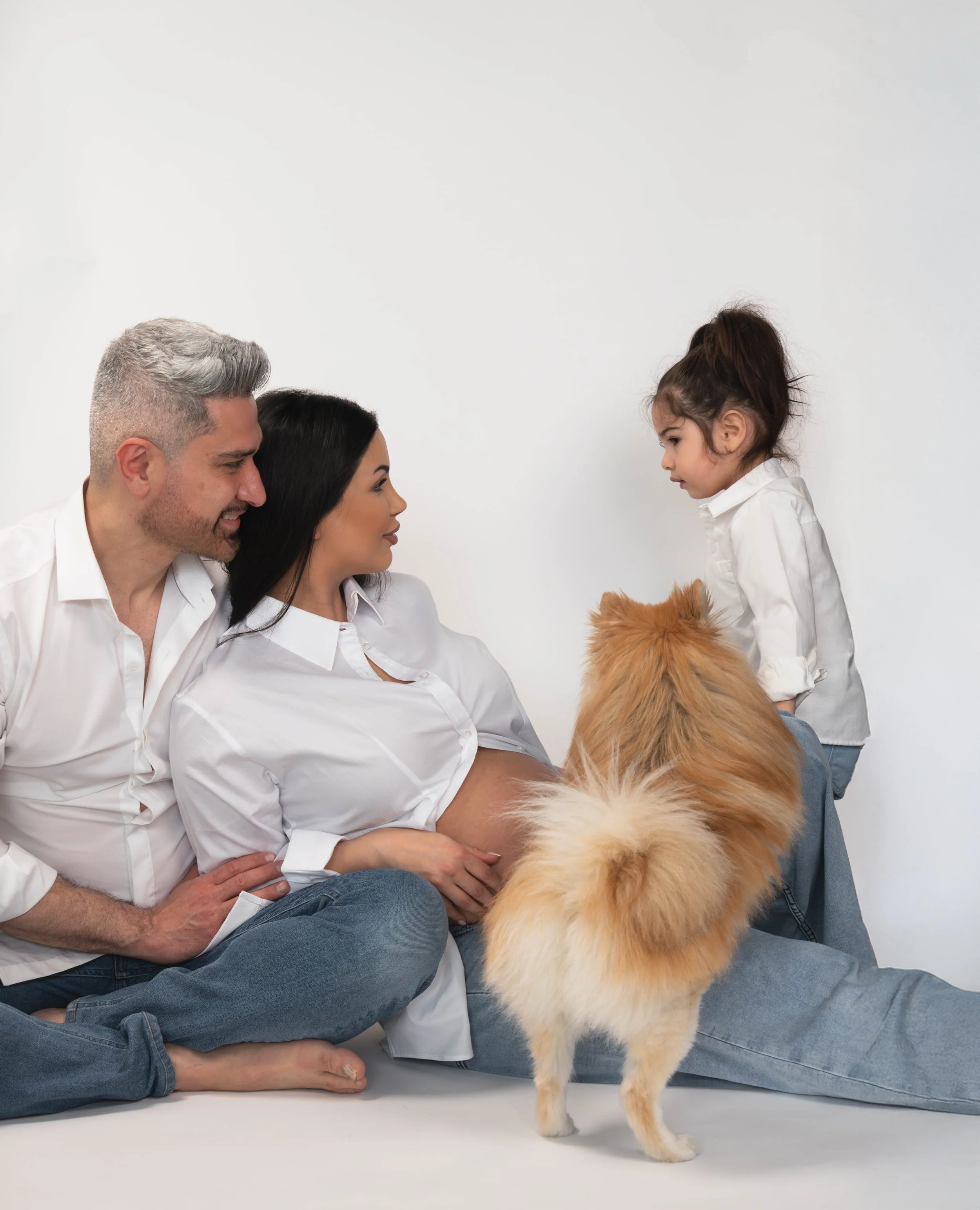 A family of three, including a man, a woman, and a young girl, sitting on the floor with a fluffy dog, all facing each other against a plain white background.