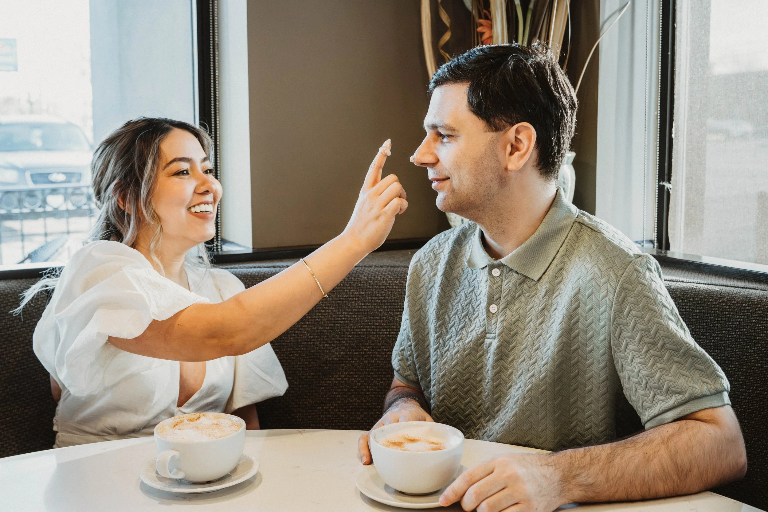 A woman playfully touching a man's nose while both sit at a table in a café, with cappuccinos in front of them.