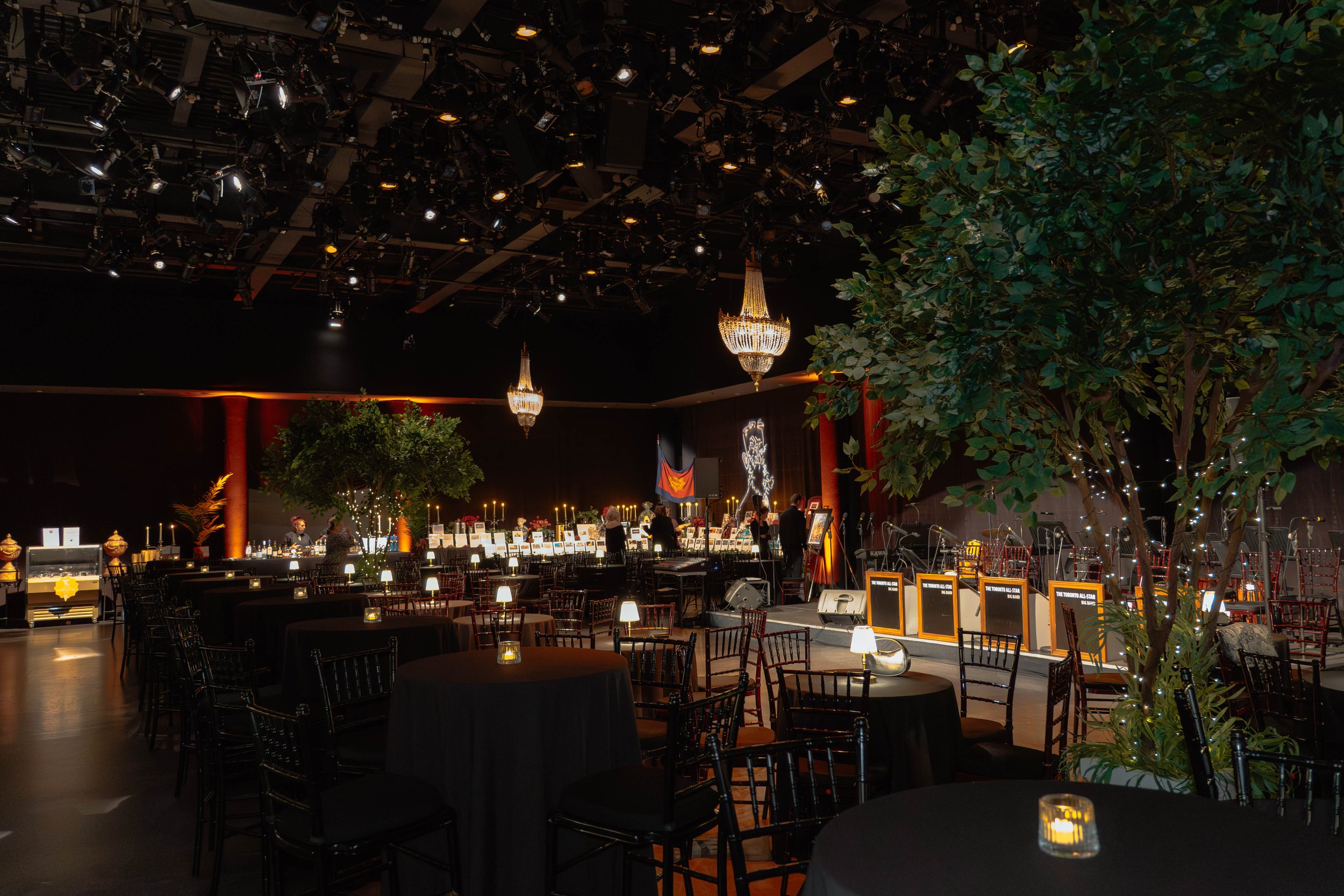 An elegant event space with round tables covered in black tablecloths, each with small candles. There are black chairs around the tables, and large chandeliers hang from the ceiling. A stage with musical instruments and microphones is set up at the f