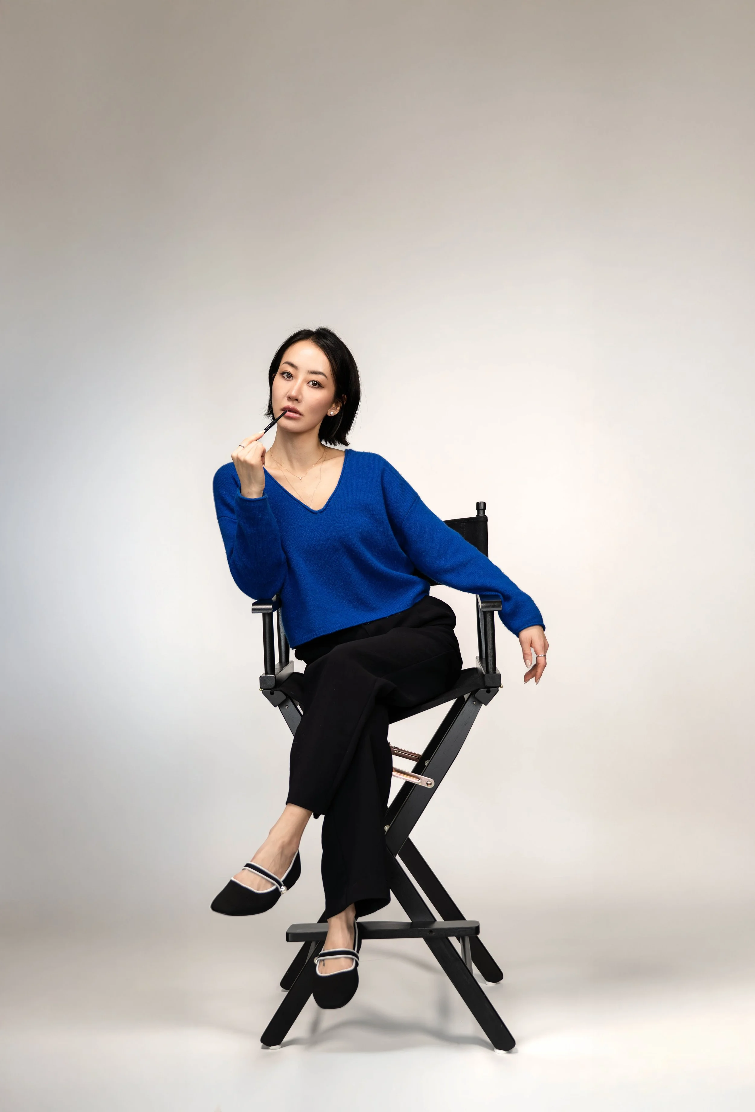 A woman with black hair sitting on a director's chair, wearing a blue sweater, black pants, and black shoes with white straps, holding a makeup brush to her lips, against a plain light-colored background.