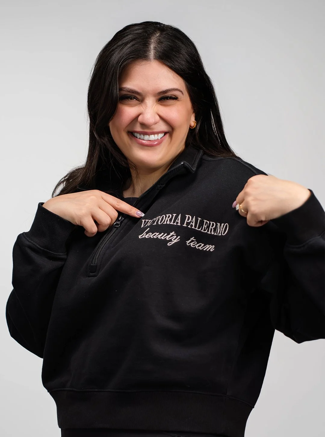 A woman with dark hair smiling and pointing to her black sweatshirt that has embroidered text reading 'VITORIA PALERMO beauty team', while flexing her right arm.