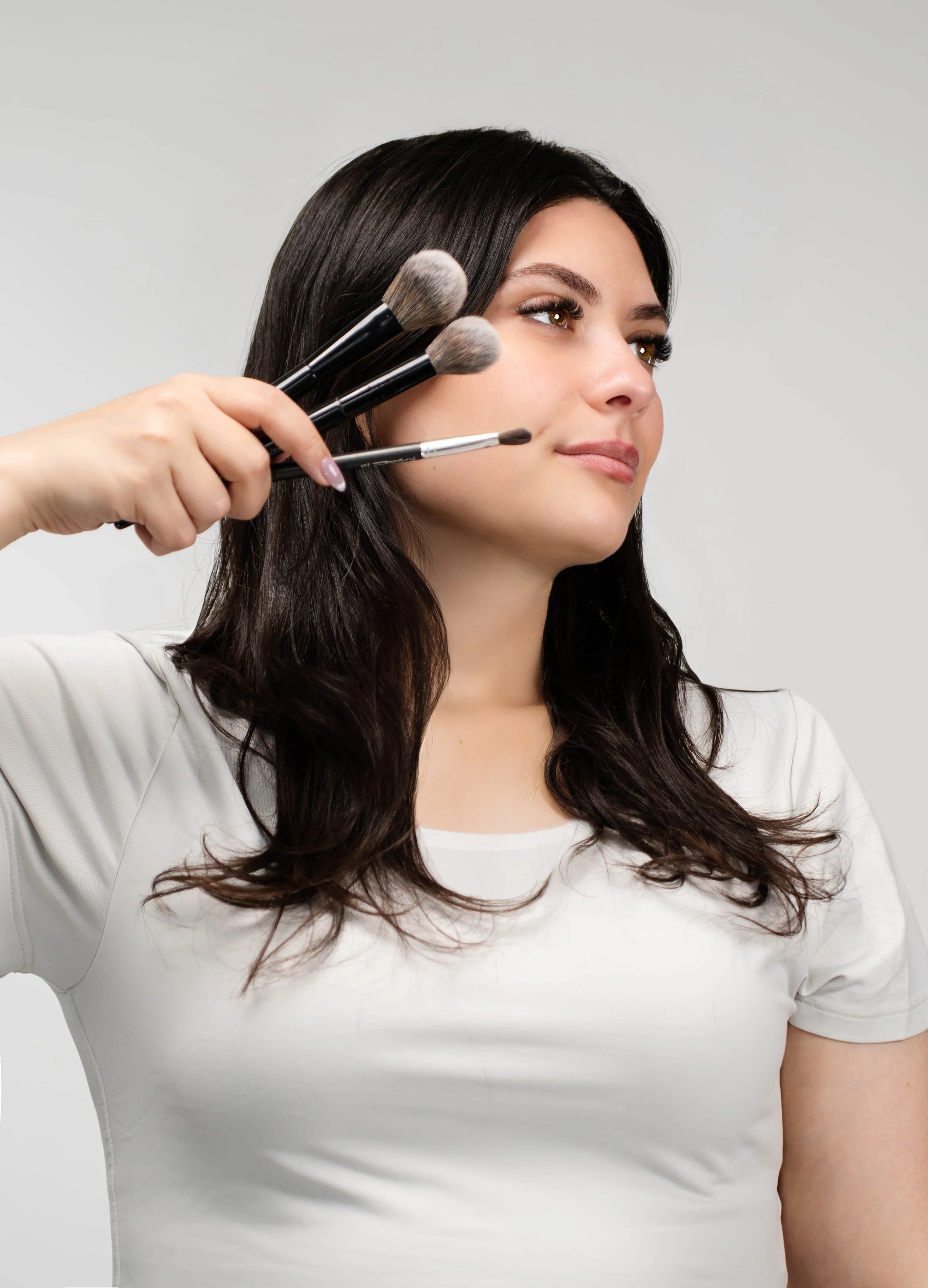 A woman with dark hair sitting calmly while a makeup artist holds four makeup brushes near her face.