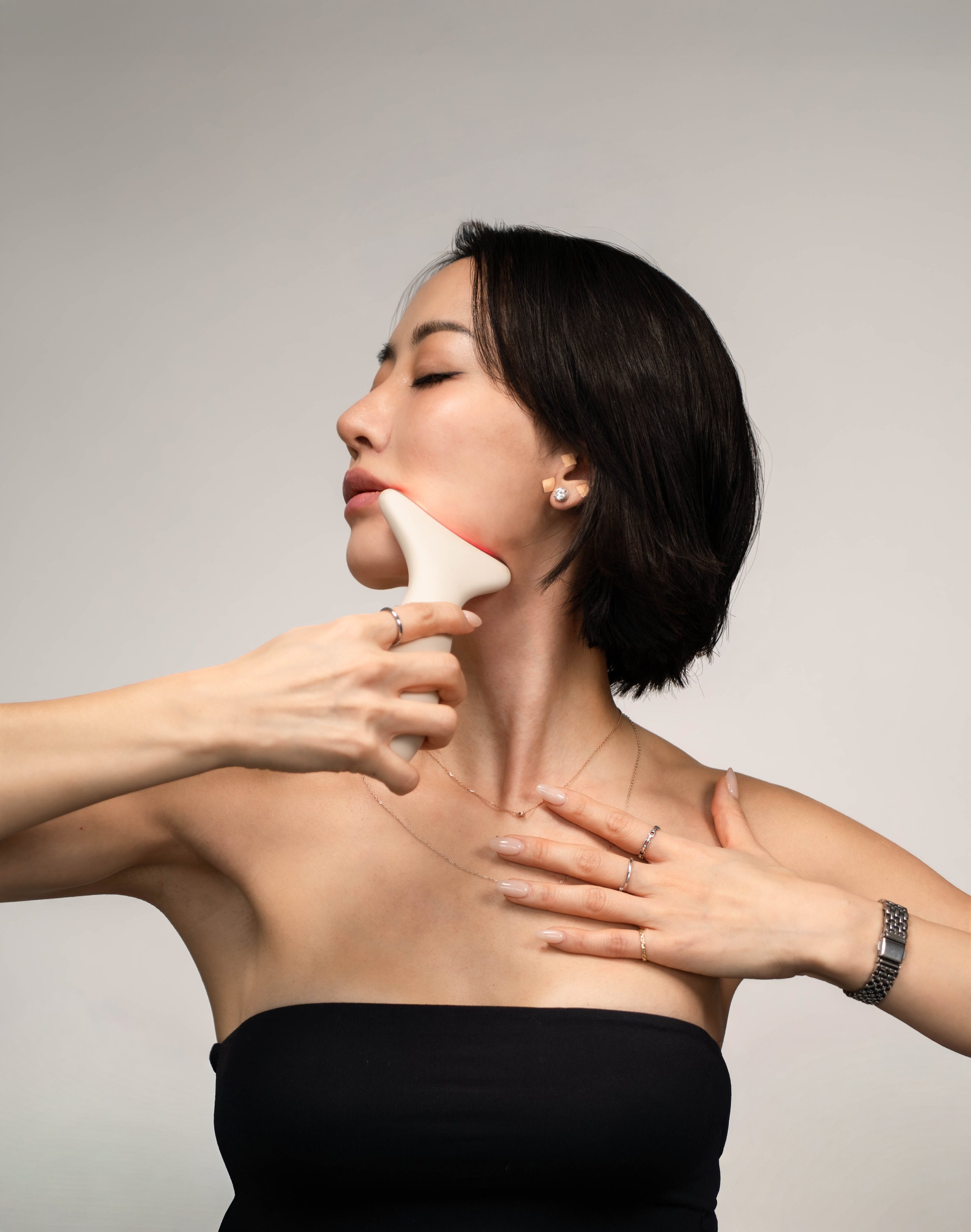 A woman with black hair getting a facial massage using a white massage tool across her jawline, with her eyes closed and her hand on her chest, wearing jewelry including rings, necklaces, and a watch.