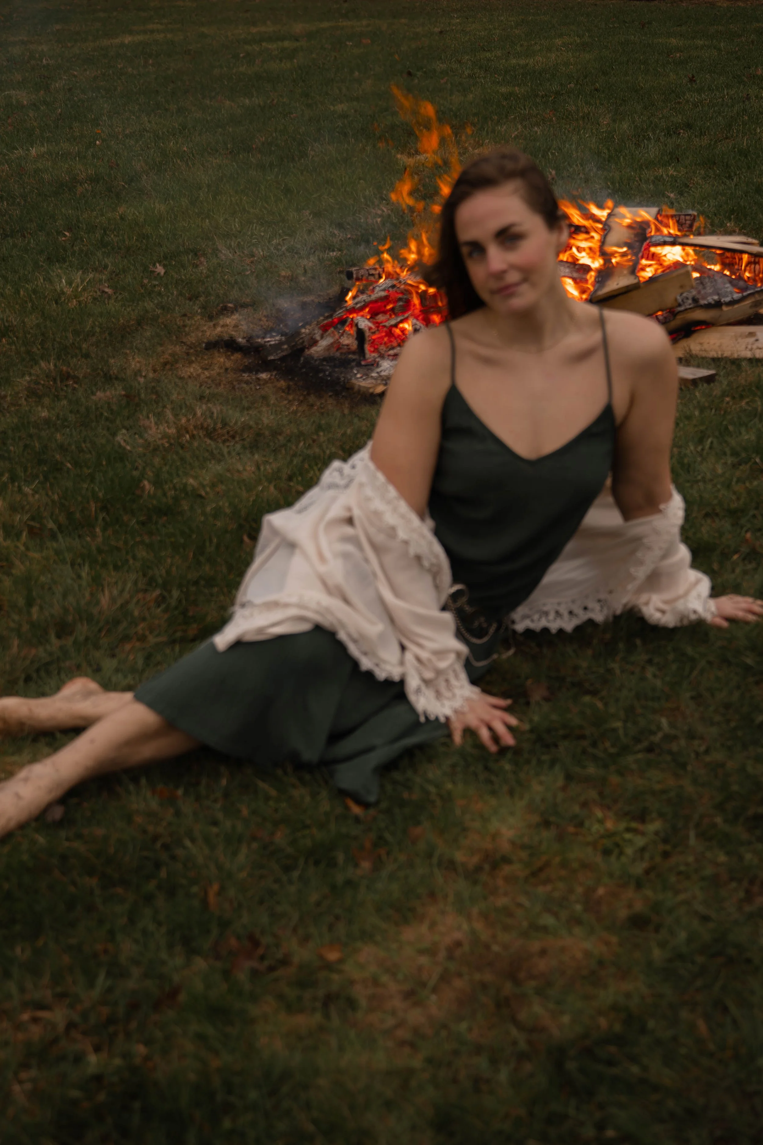 A woman in a dark green slip dress and a lace-trimmed cream shawl sitting on grass in front of a fire with burning logs, during dusk or evening.