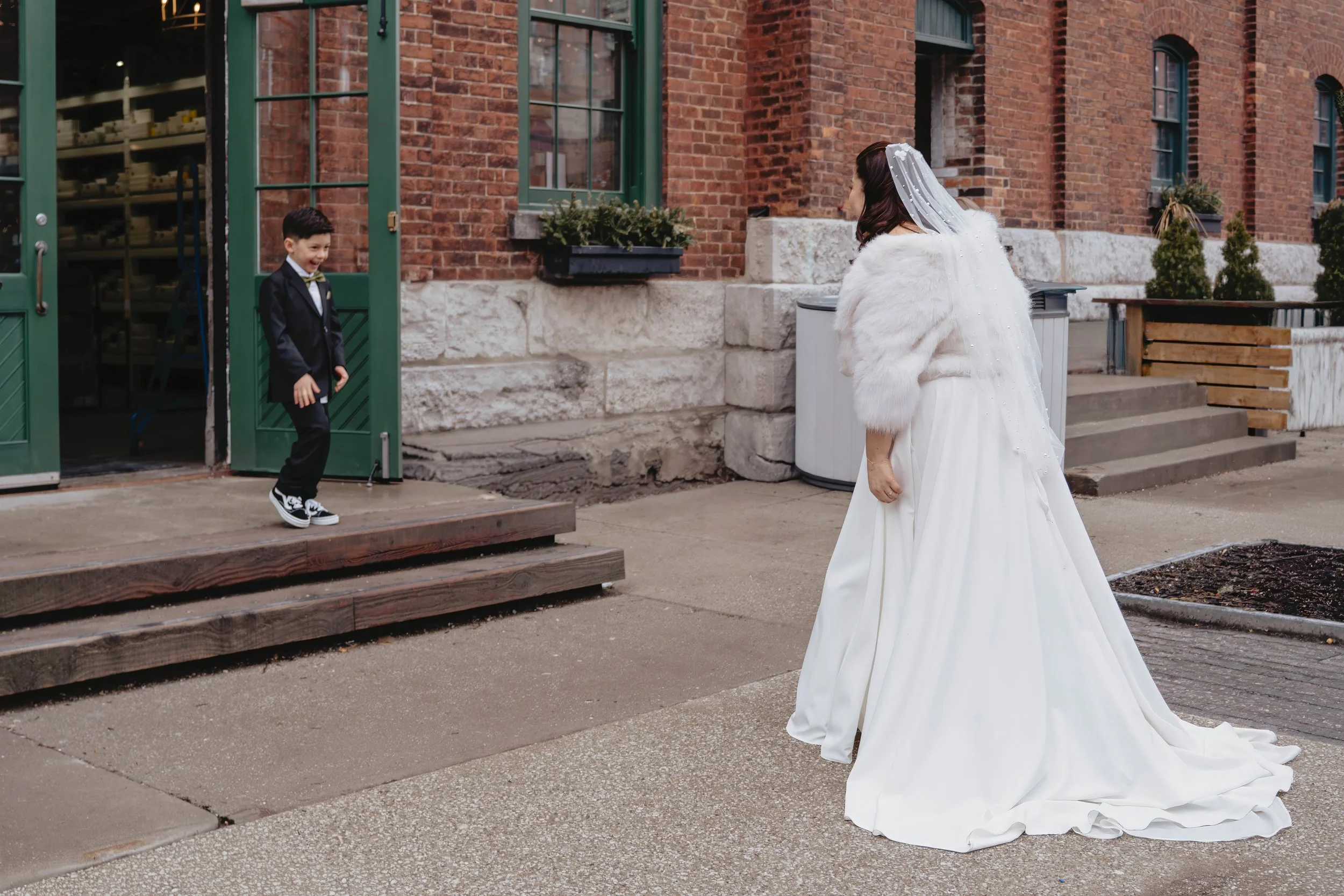 A young boy dressed in a black suit and sneakers is standing on a small wooden platform, smiling, as he sees a bride in a white wedding dress and veil, wearing a white fur stole, approaching her outside in front of a brick building.