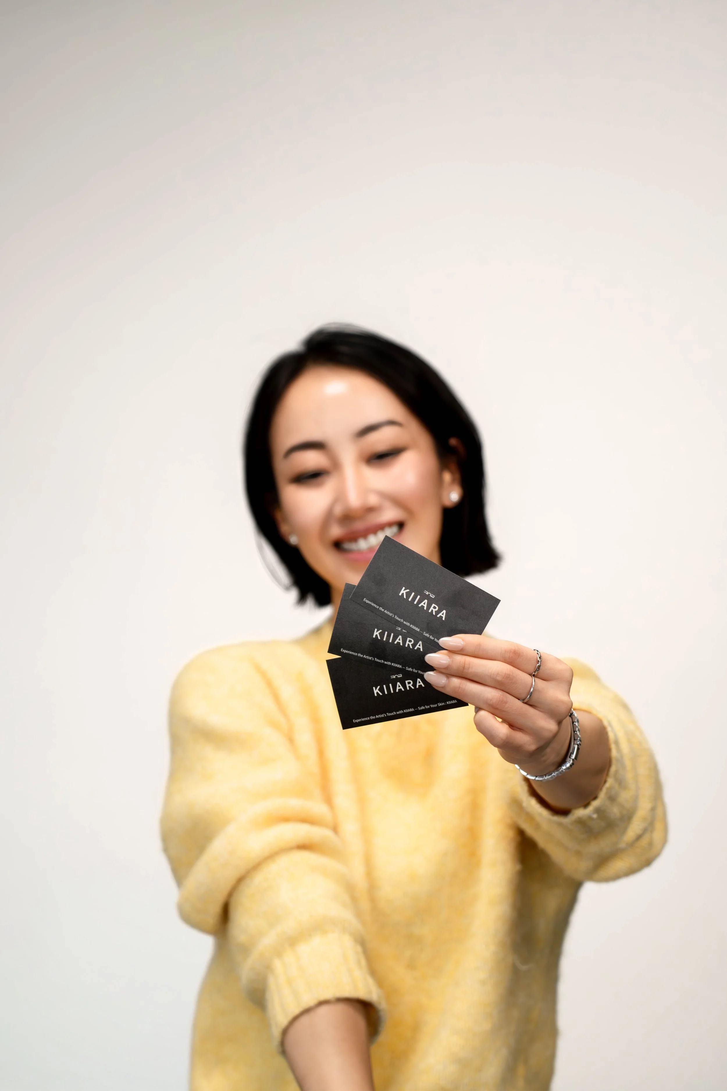 A smiling woman in a yellow sweater holding up three black business cards with the text "KIIARA" on them.