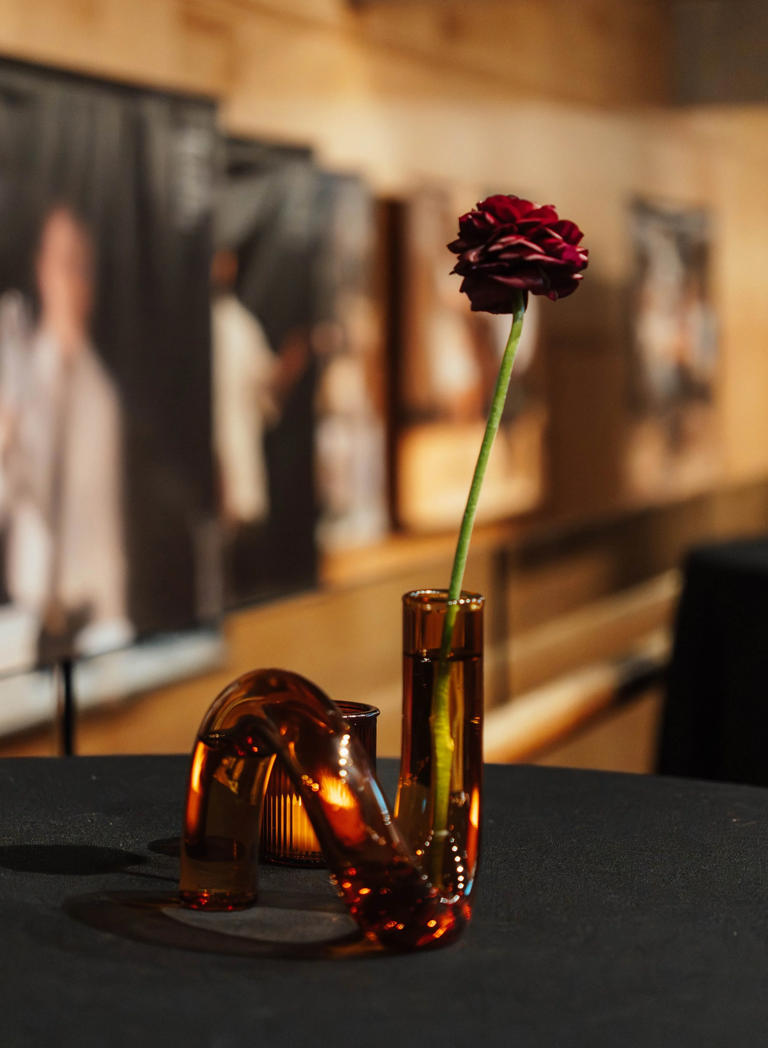 A dark red flower in a tall, narrow amber vase, with a smaller amber glass sculpture beside it, on a black table with a blurred background of framed pictures or artwork.