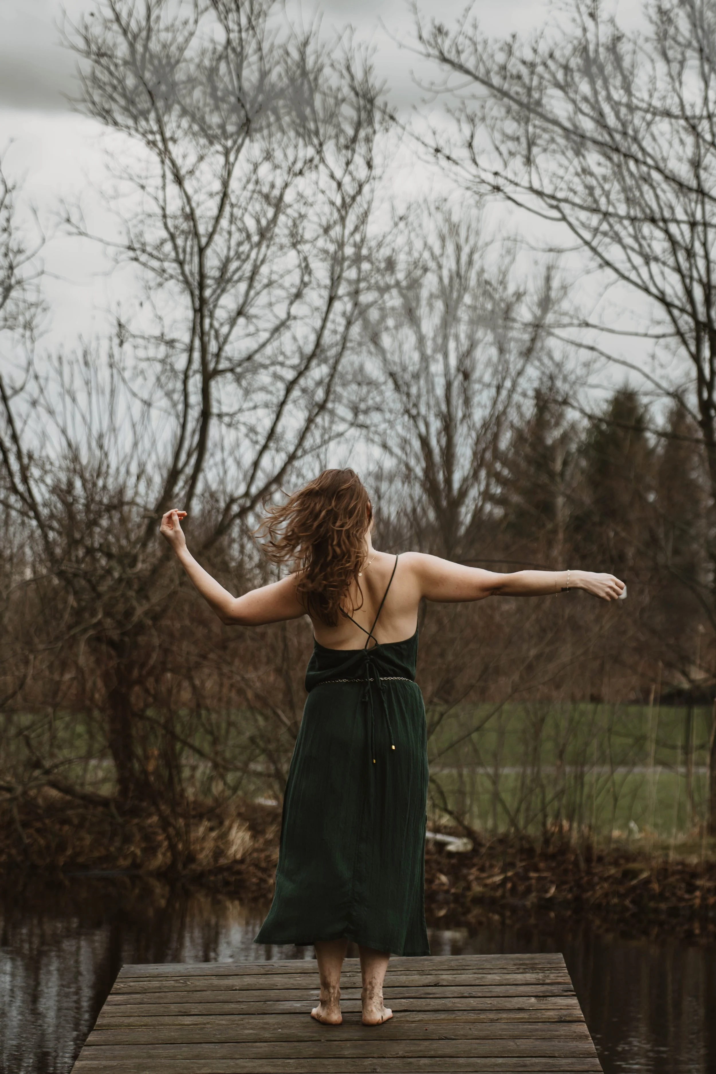 Woman with long curly hair in a black dress standing on a wooden dock with arms outstretched near a body of water, surrounded by leafless trees on a cloudy day.