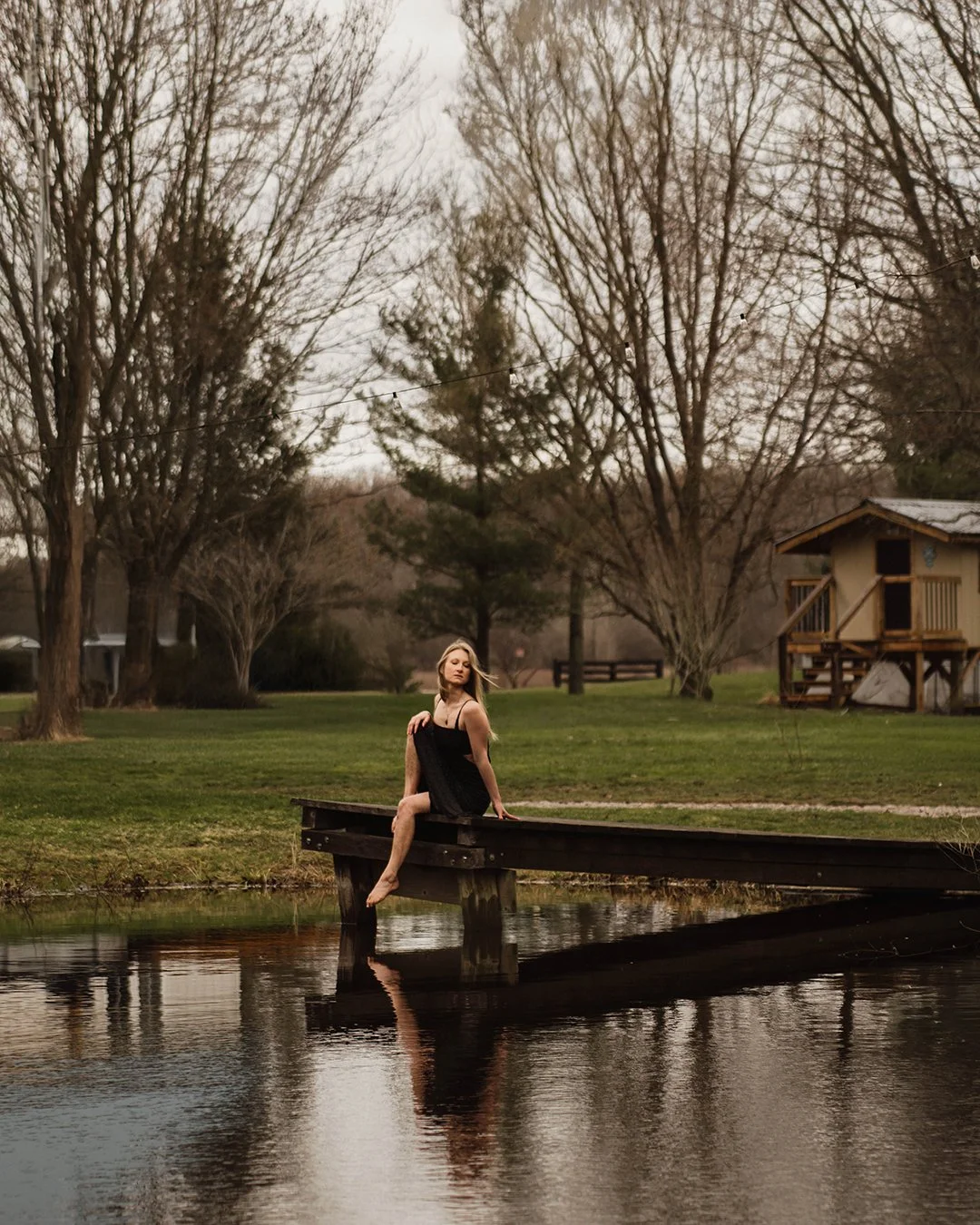 A woman in a black dress sitting on the edge of a small wooden dock by a pond, surrounded by leafless trees, a grassy lawn, and a small wooden house in the background, during overcast weather.