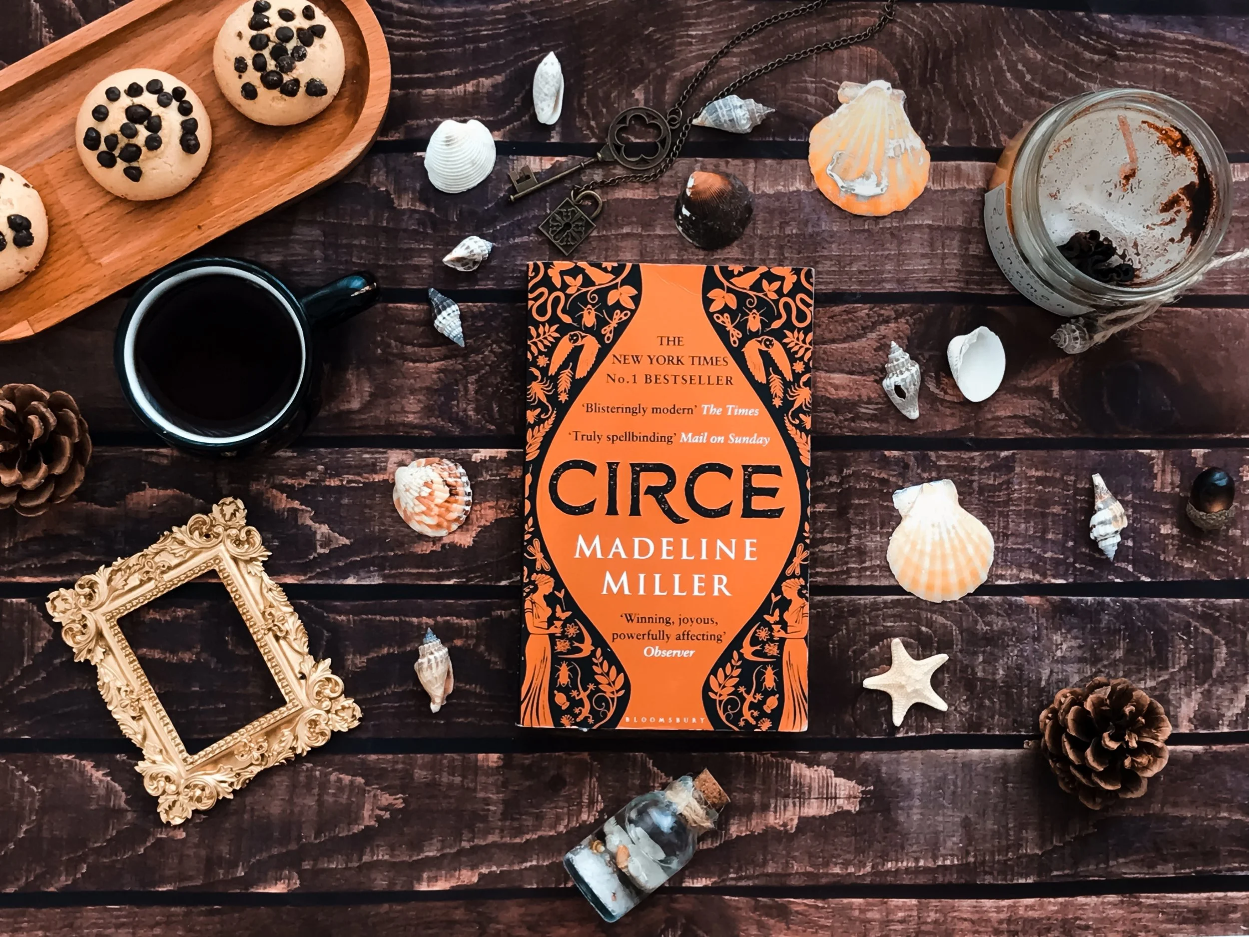 A flat lay of a book titled 'Circe' by Madeline Miller on a dark wooden surface, surrounded by seashells, pinecones, a cup of coffee, a jar with remnants of a dark beverage, an ornate empty picture frame, a small bottle of what appears to be salt, a 