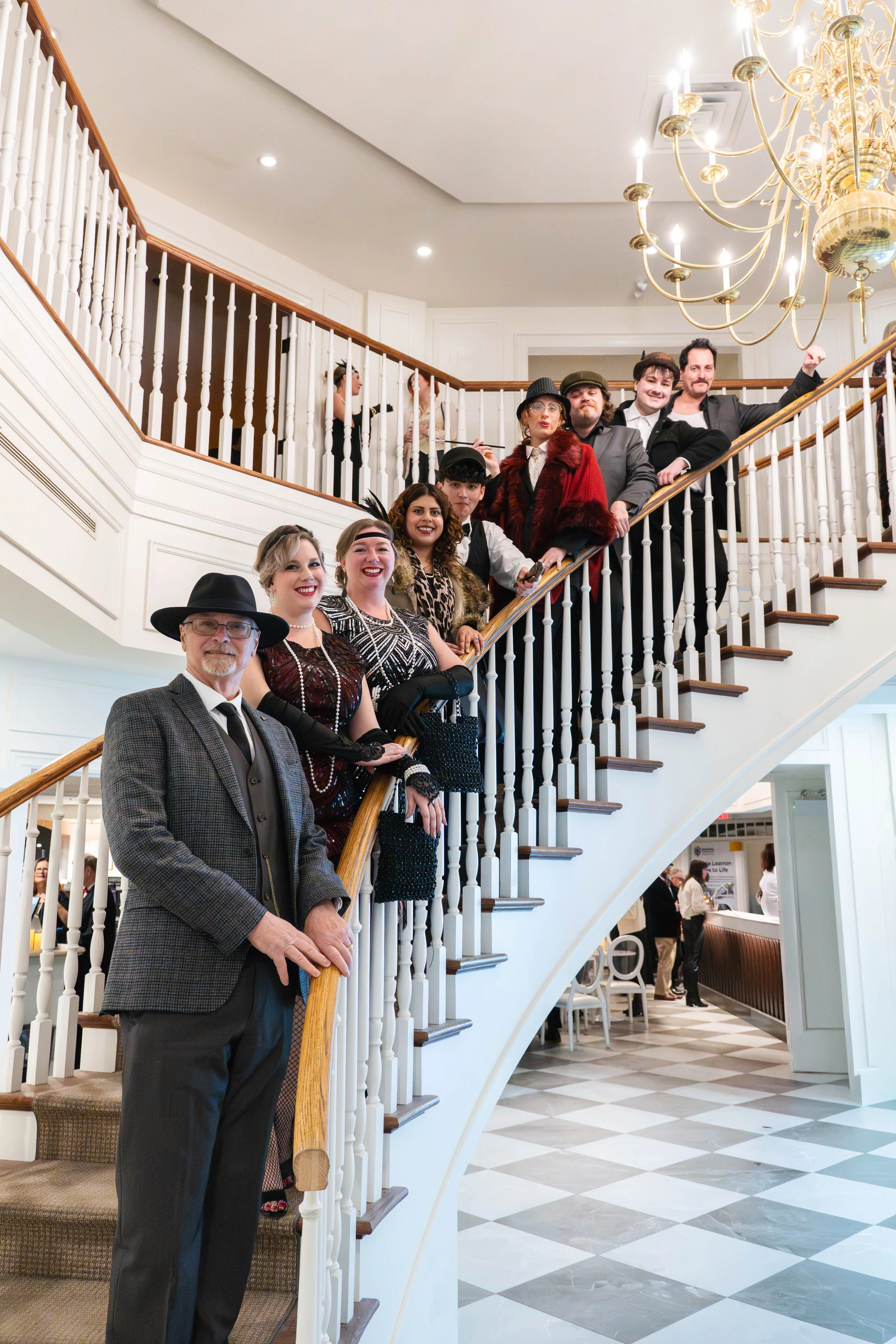 Group of people dressed in vintage or retro clothing standing on a curved staircase inside a well-lit interior with a chandelier overhead.