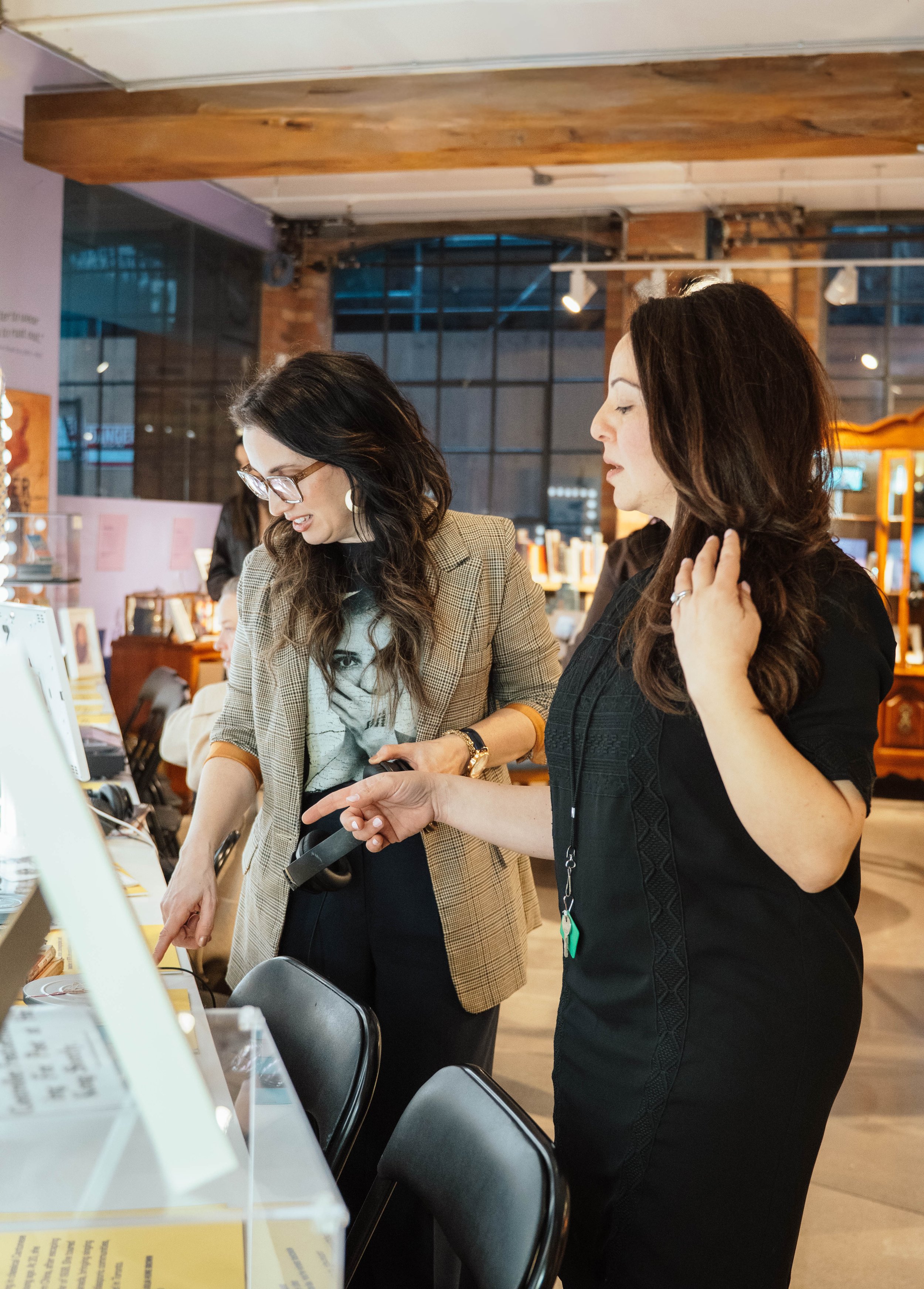 Two women look at products on display in a store, one pointing at an item while the other observes, with shelves of books and displays behind them.