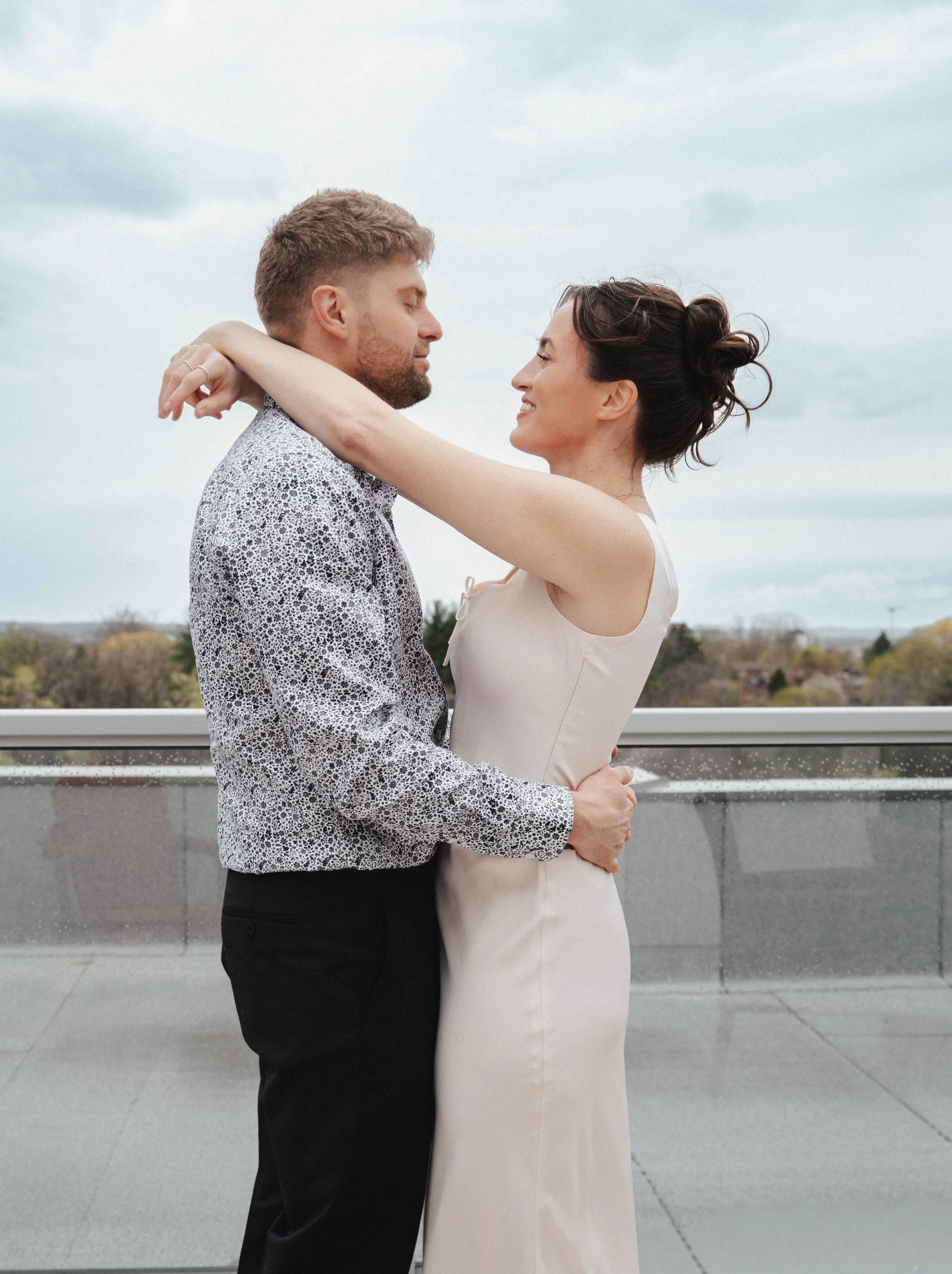 A man and a woman are embracing each other on a balcony during the daytime. The man has short, light brown hair and a beard, and the woman has dark hair styled in an updo. They are standing close, smiling with eyes closed, and the woman has her arms 