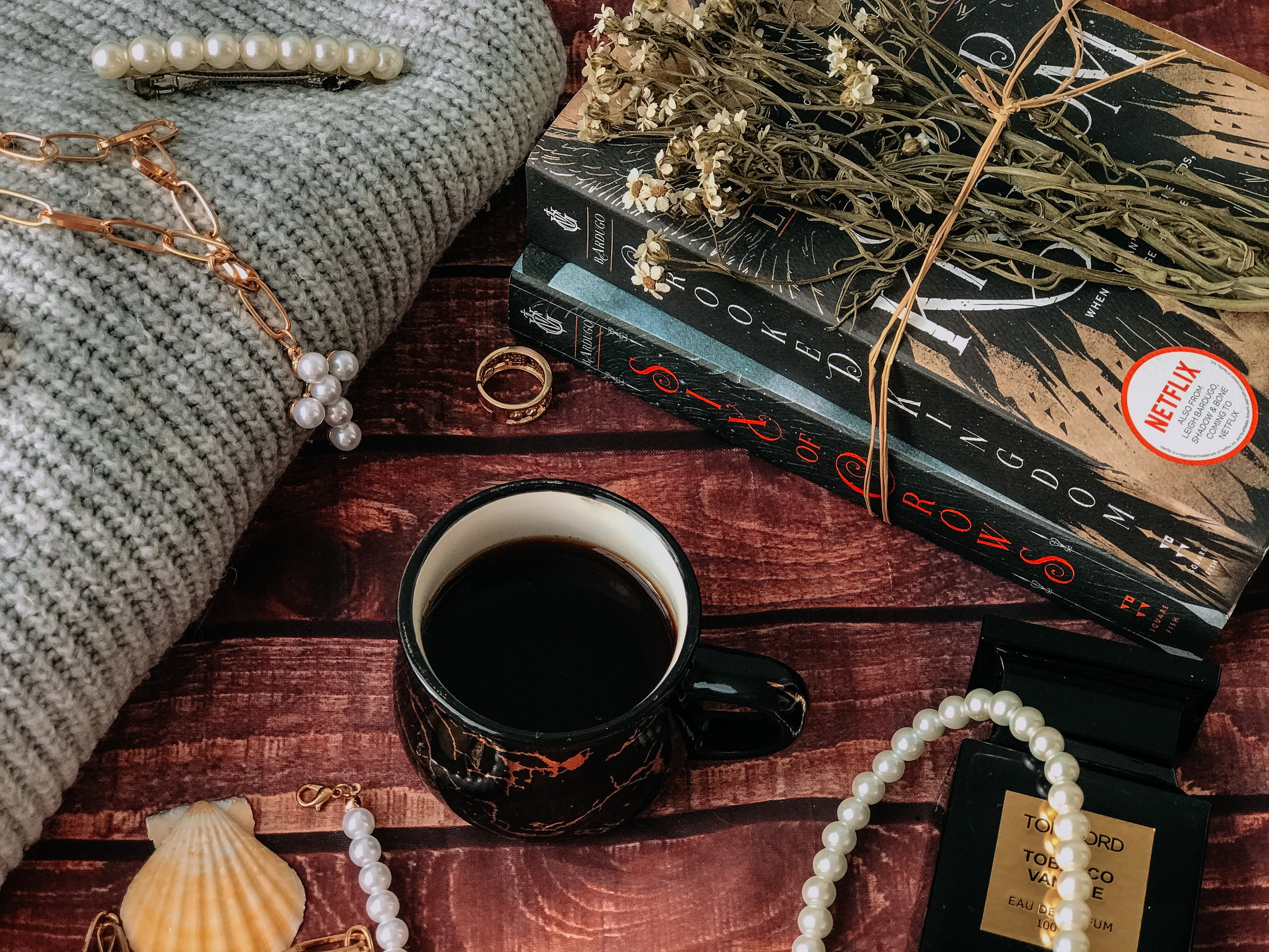 A flat lay of a cozy scene with a gray sweater, pearl necklaces, and a gold ring, alongside a mug of coffee, a shell, books, a bouquet of dried flowers, and a black perfume box on a wooden surface.
