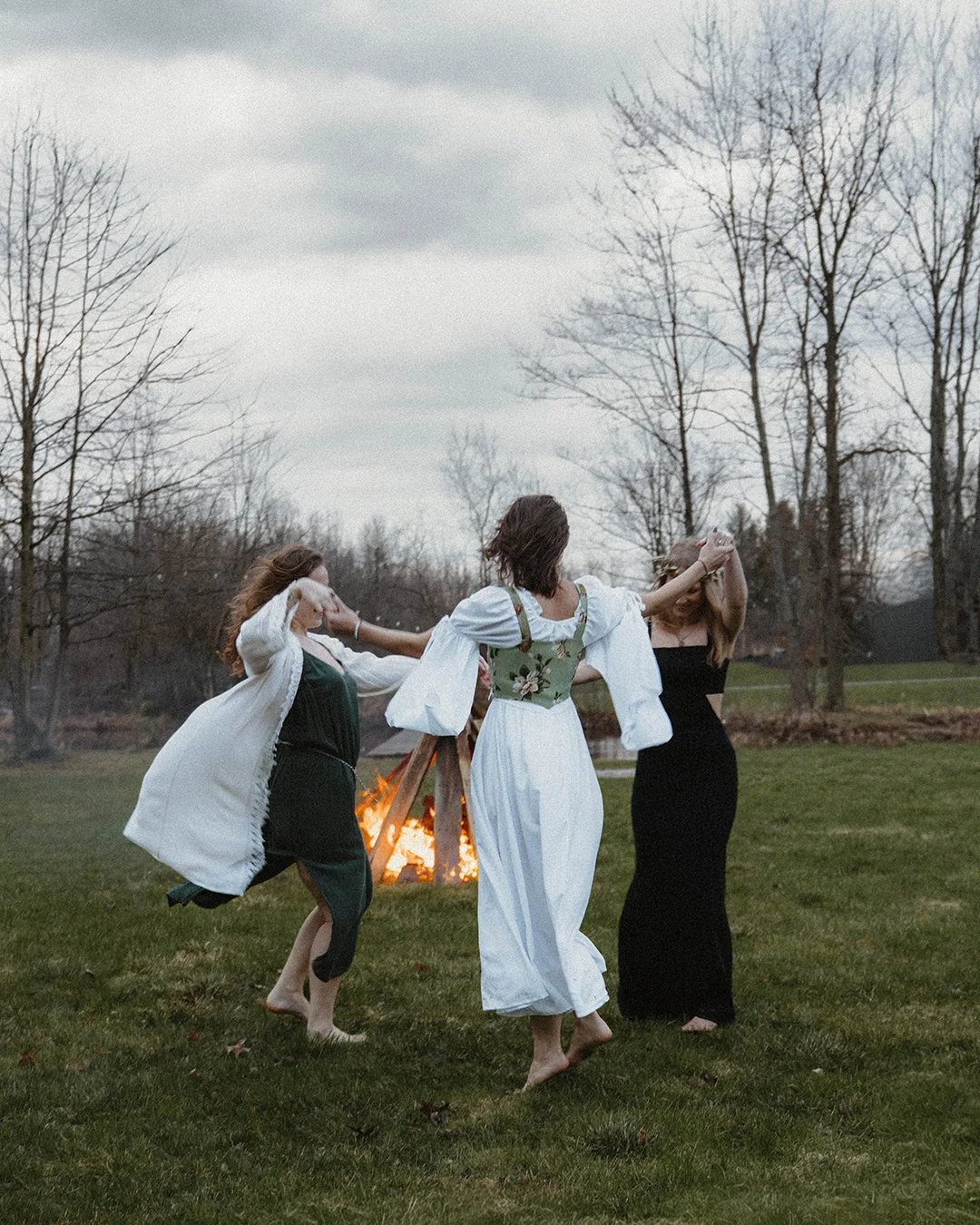 Three women dancing in a grassy yard near a bonfire on a cloudy day, with leafless trees in the background.