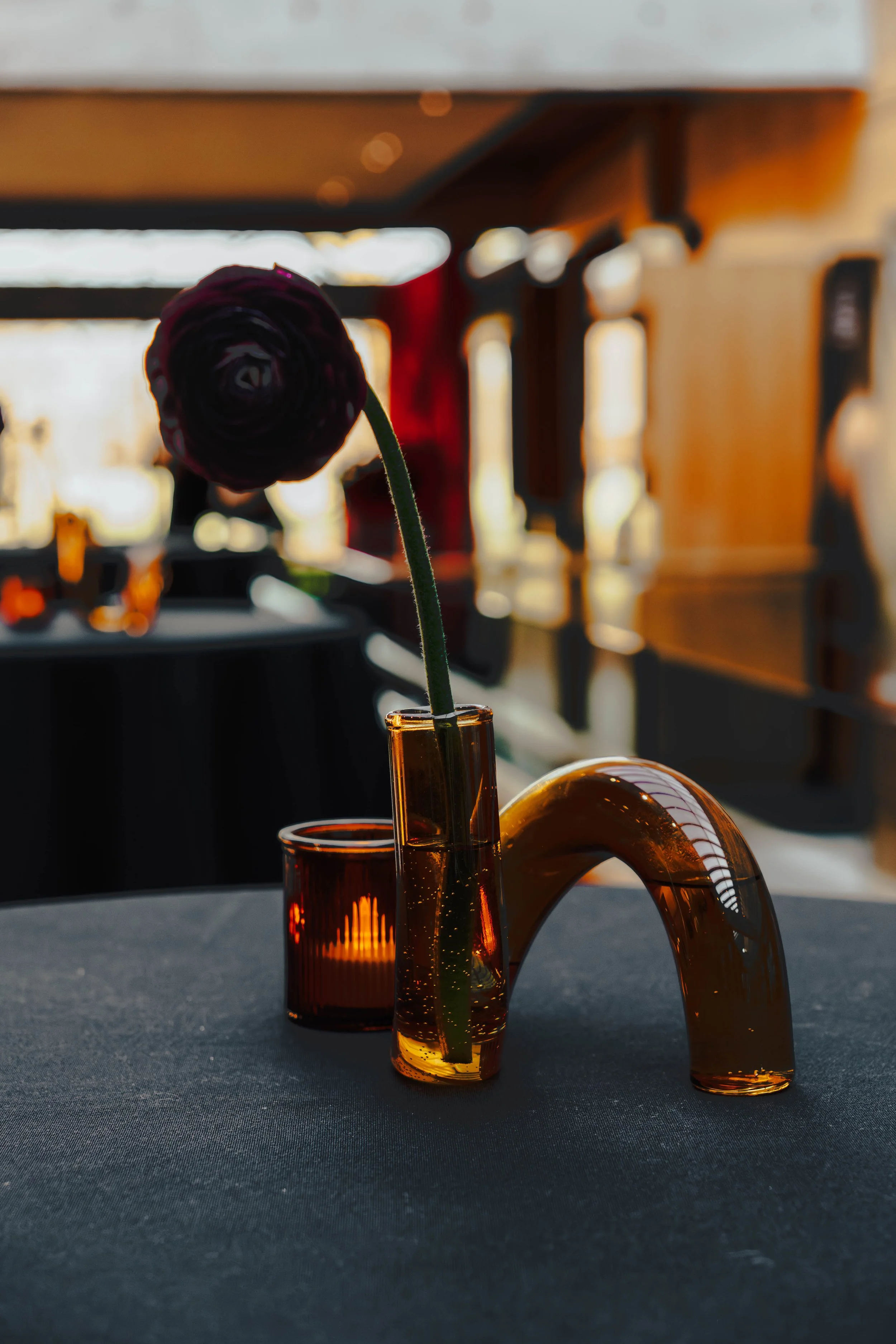 A dark purple flower in a tall amber glass vase on a black table, with a small amber candle holder nearby, in a warmly lit interior space.