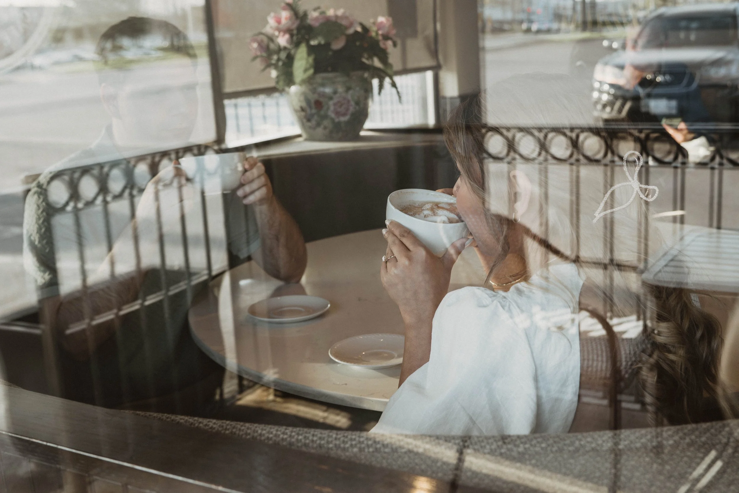 A woman with long hair holding a steaming cup of coffee inside a cafe, viewed through the window with reflections of the street and a car outside.