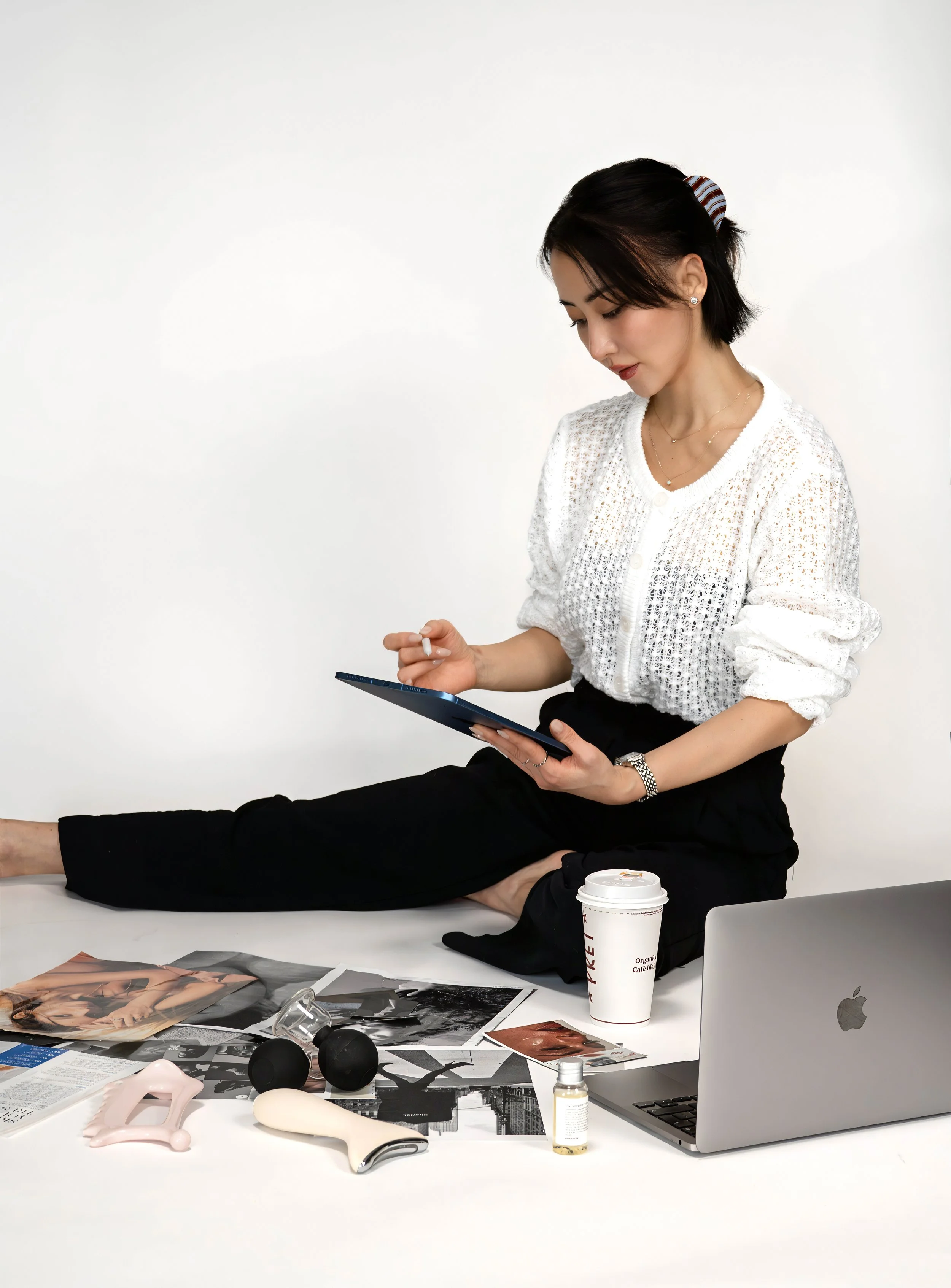 A woman sitting cross-legged on the floor, surrounded by magazines, a laptop, a coffee cup, a bottle, and various objects, looking at a digital tablet.