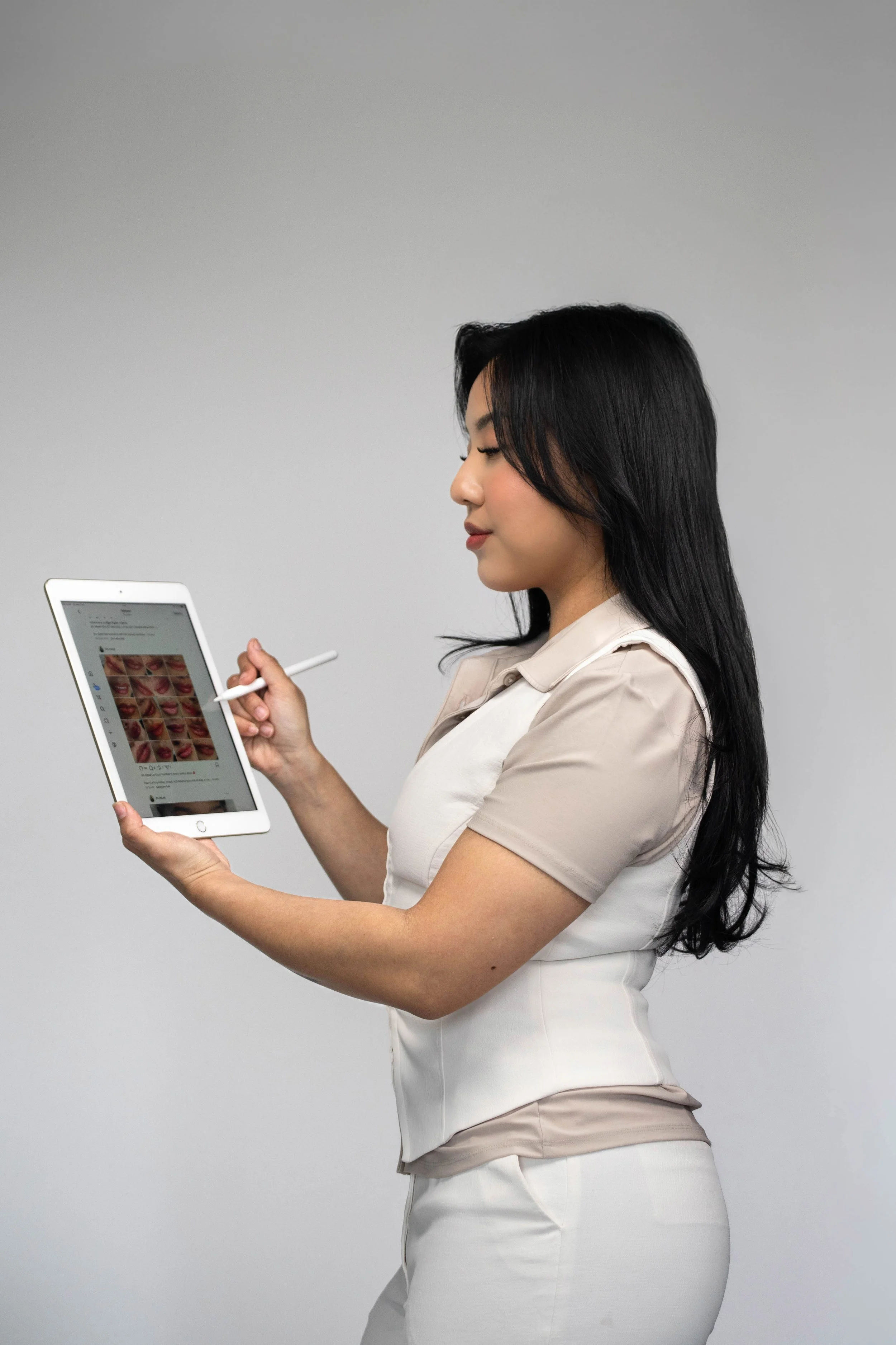 A woman with long black hair in profile, dressed in a beige and white outfit, using a white stylus to select images on a digital tablet against a plain gray background.