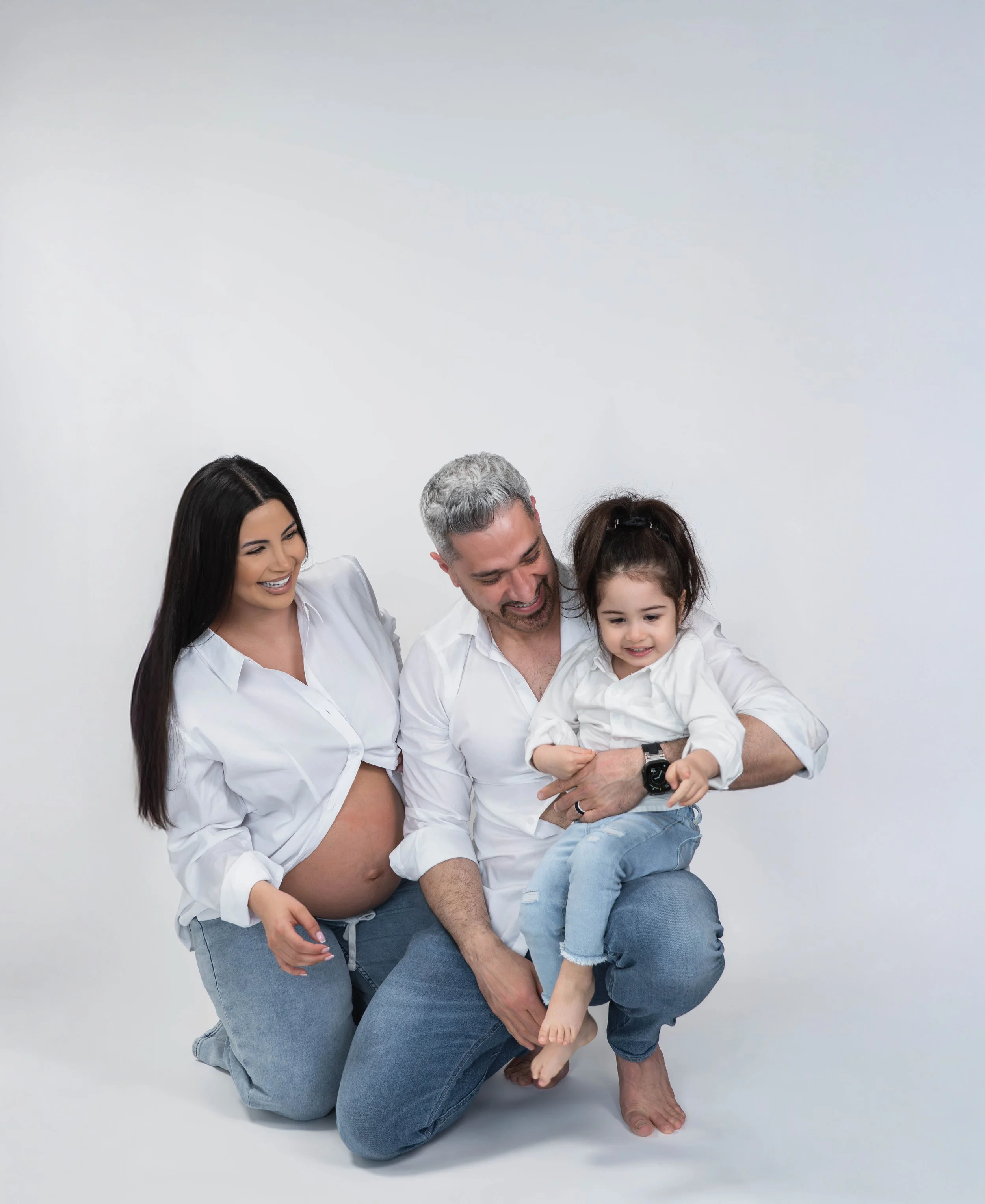 A multigenerational family of five, including a man, a woman, and a young girl, all smiling and laughing together, with the man holding the girl on his lap against a plain white background.
