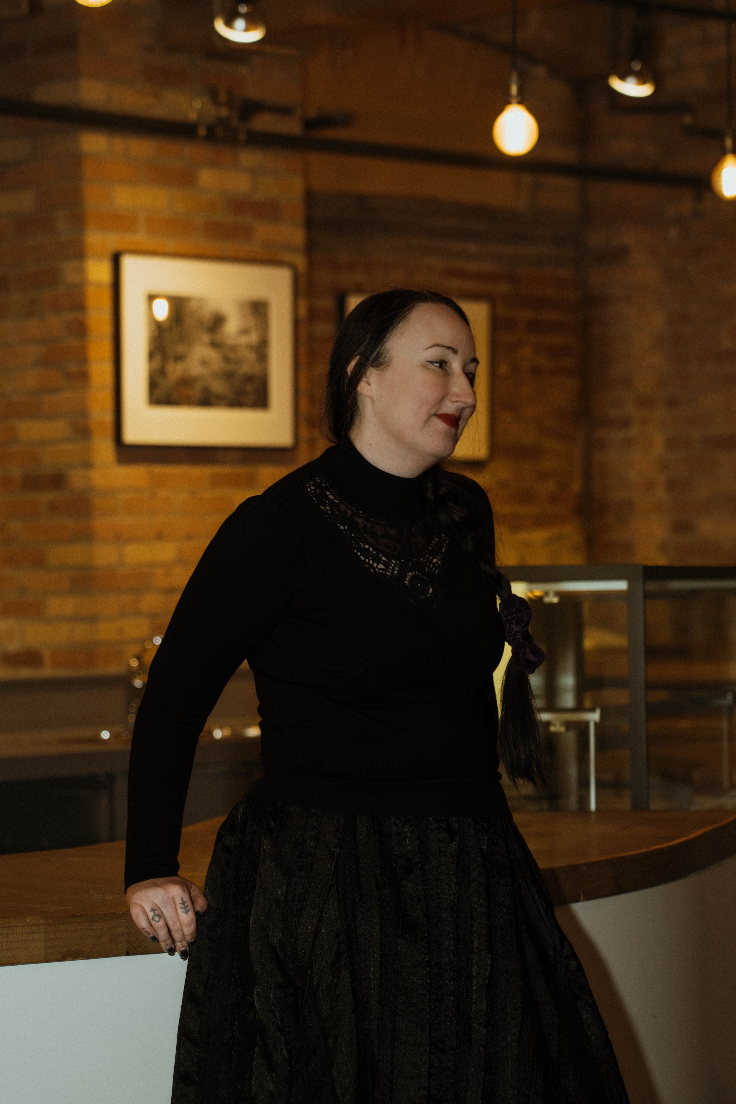 A woman with dark hair in a braid, wearing a black turtleneck and skirt, standing indoors against a brick wall with framed artwork and hanging lights.