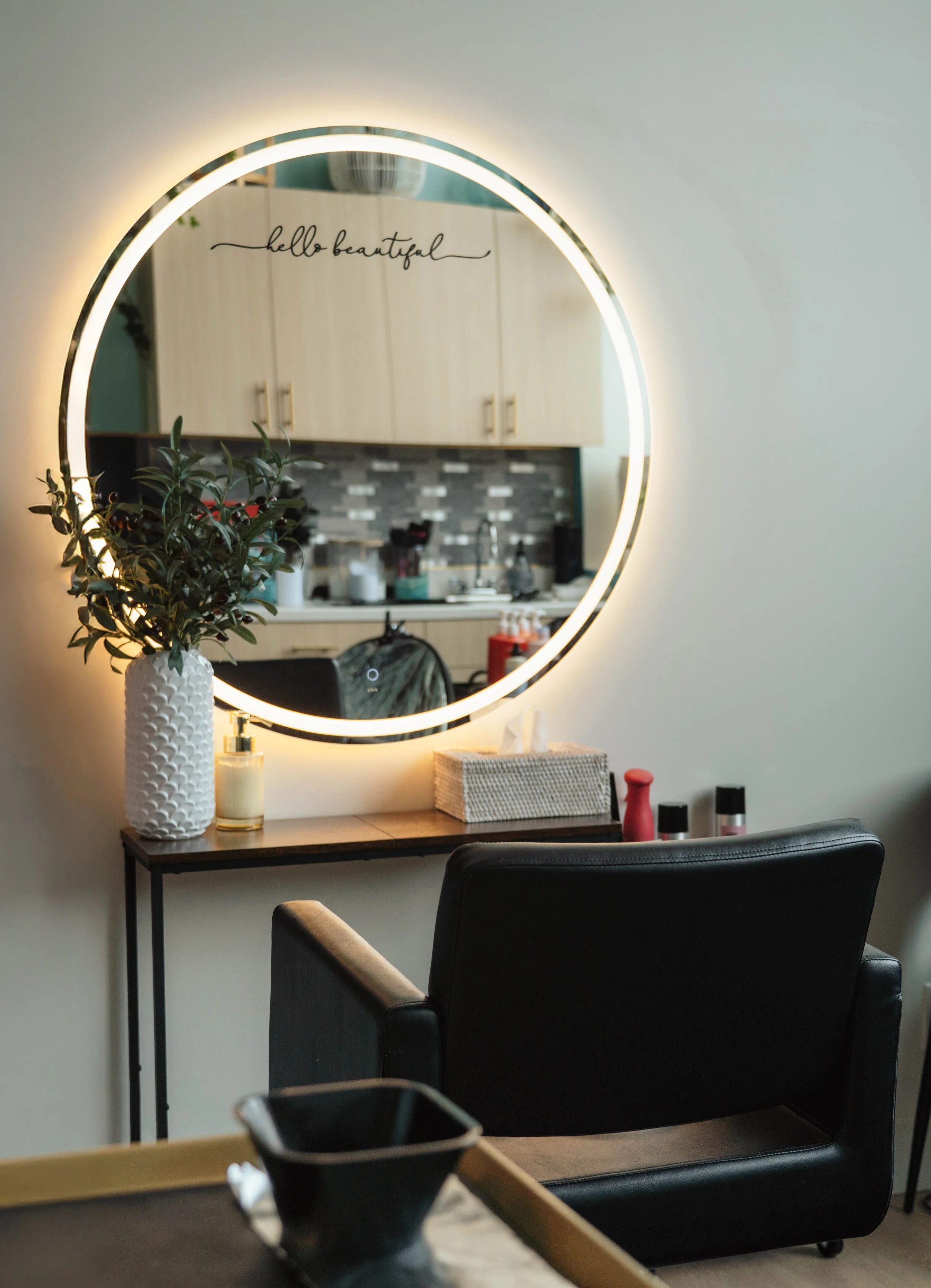 A round illuminated mirror above a wooden table with a white textured vase holding green foliage, a bottle of lotion, tissue box, and small bottles. A black chair is in front of the table, and the reflection shows a kitchen with cabinets, tiles, and 