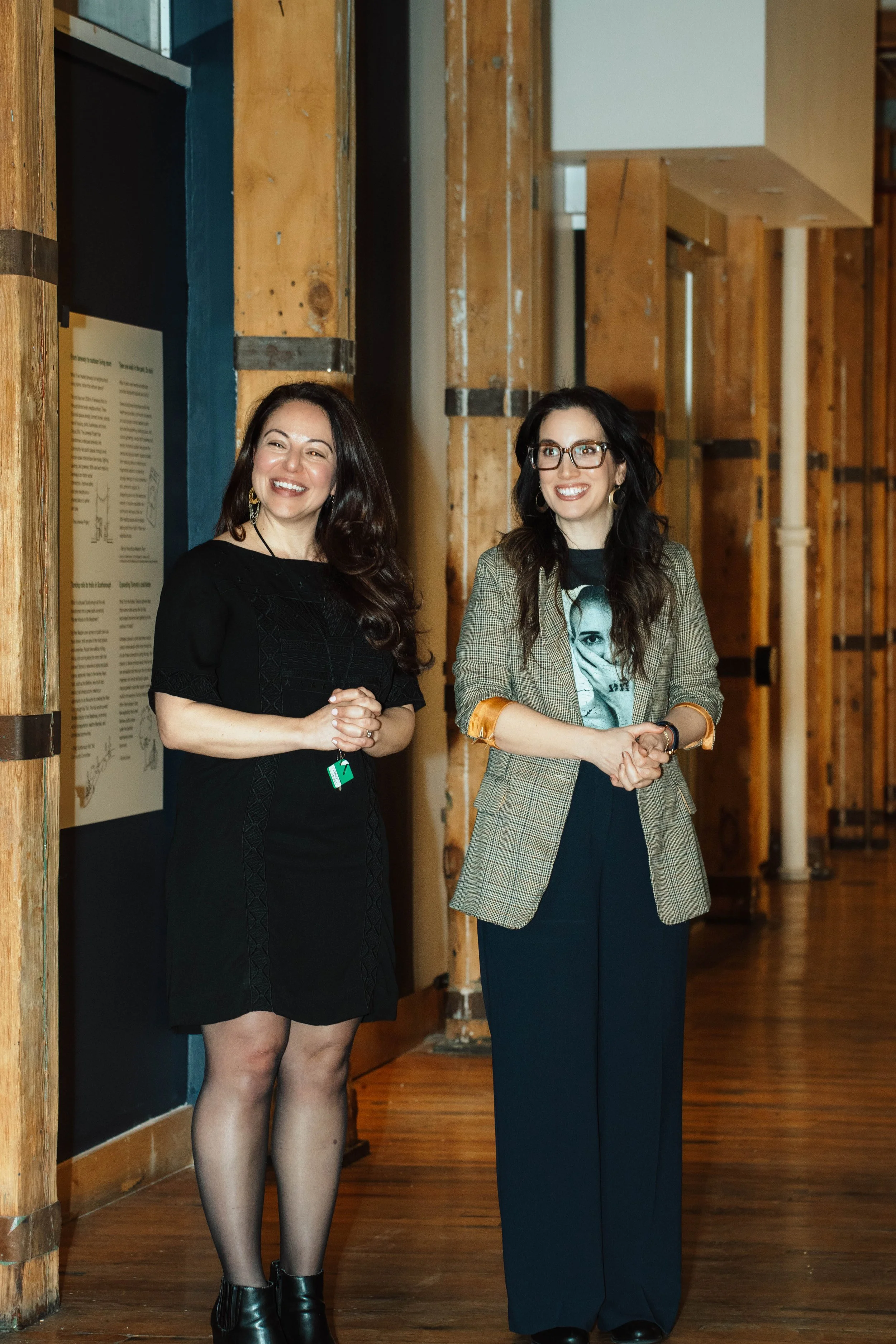 Two women are standing in a room with wooden walls, smiling and talking. One woman is wearing a black dress and has long dark hair. The other woman is wearing a plaid blazer, glasses, and has long dark hair.