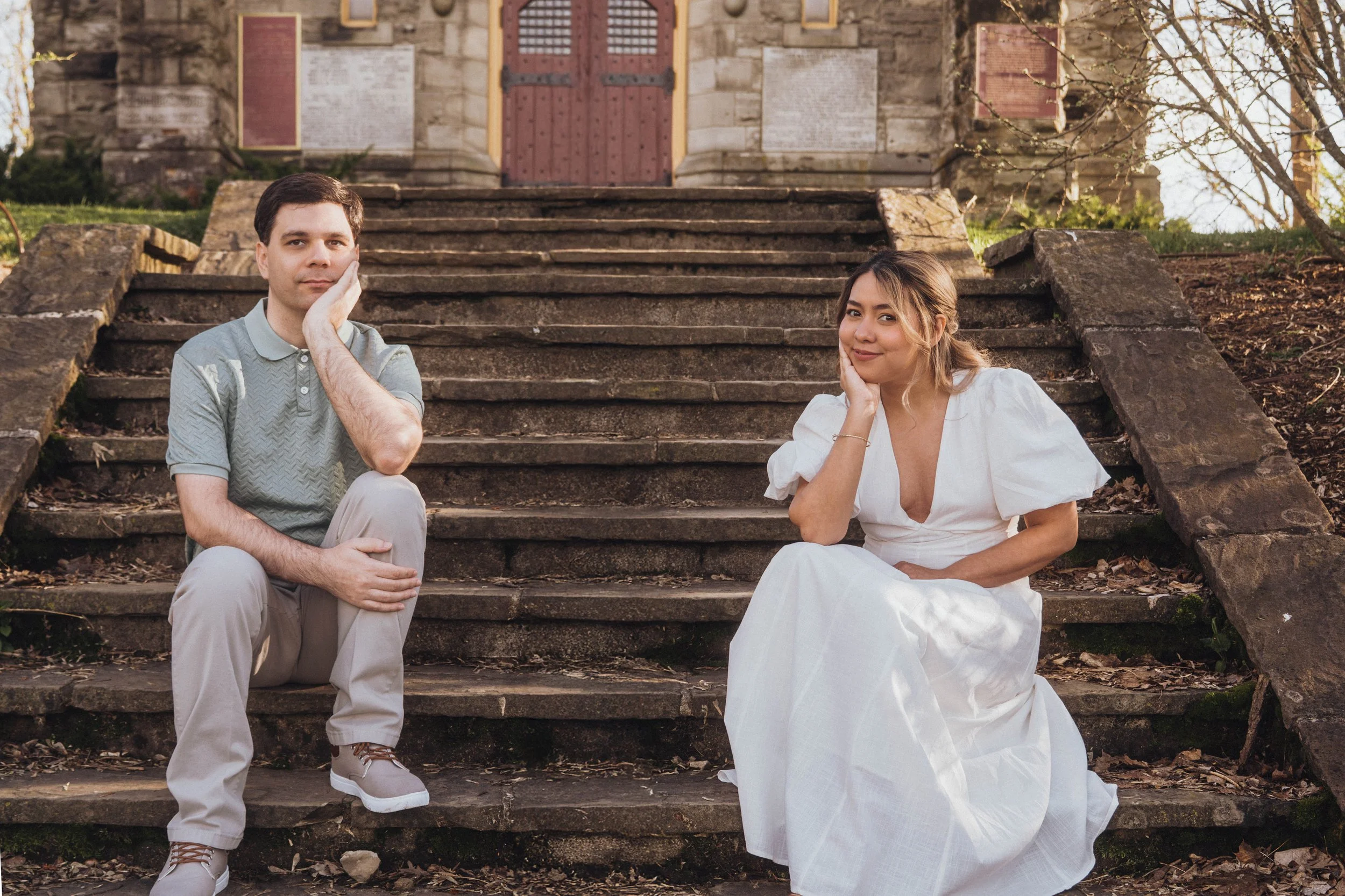 A man and woman seated on stone steps in front of a church, with the woman wearing a white dress and the man in a gray polo shirt, both resting their heads on their hands.