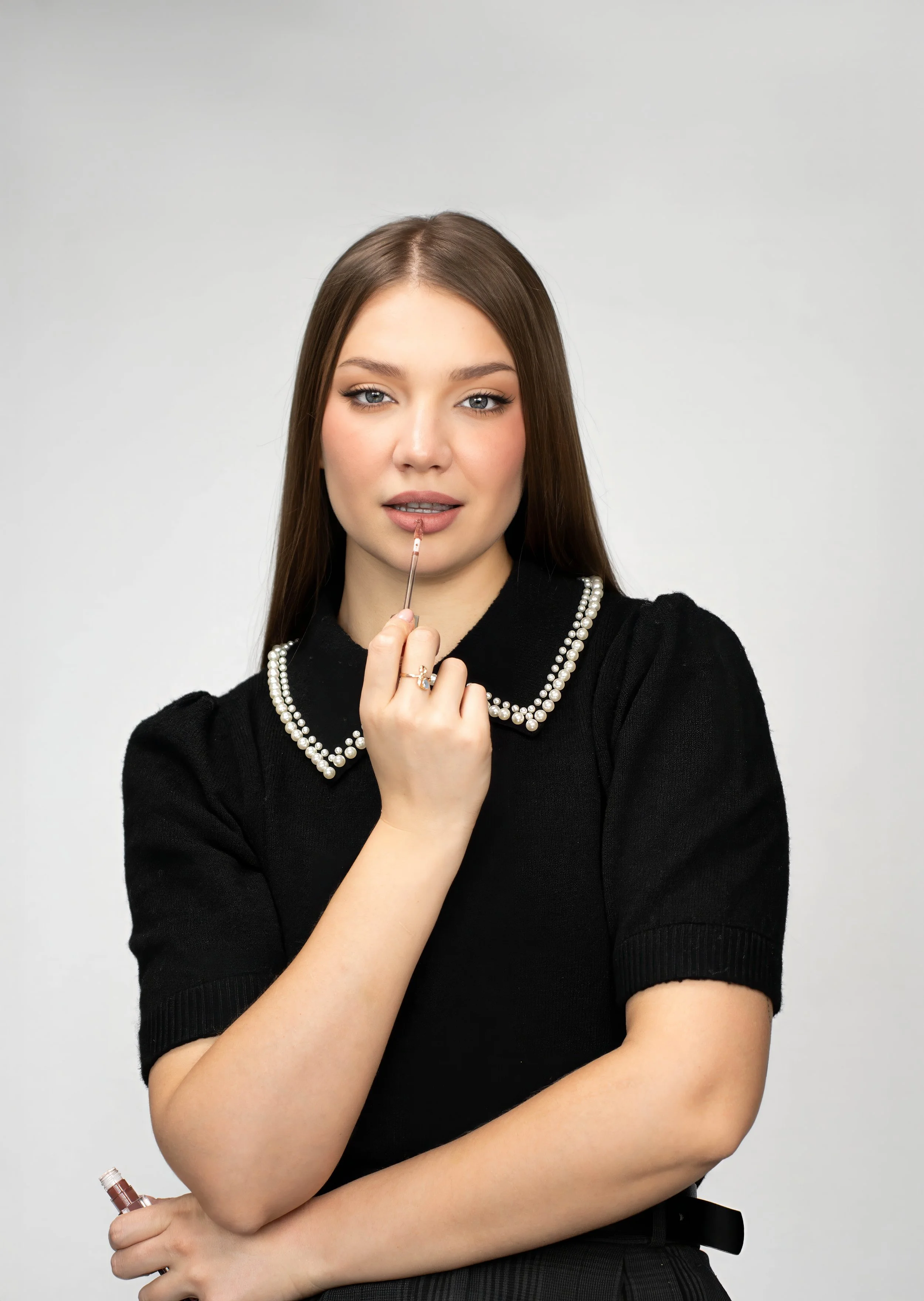 Young woman with long brown hair applying lip gloss on her lips in front of a plain light gray background.
