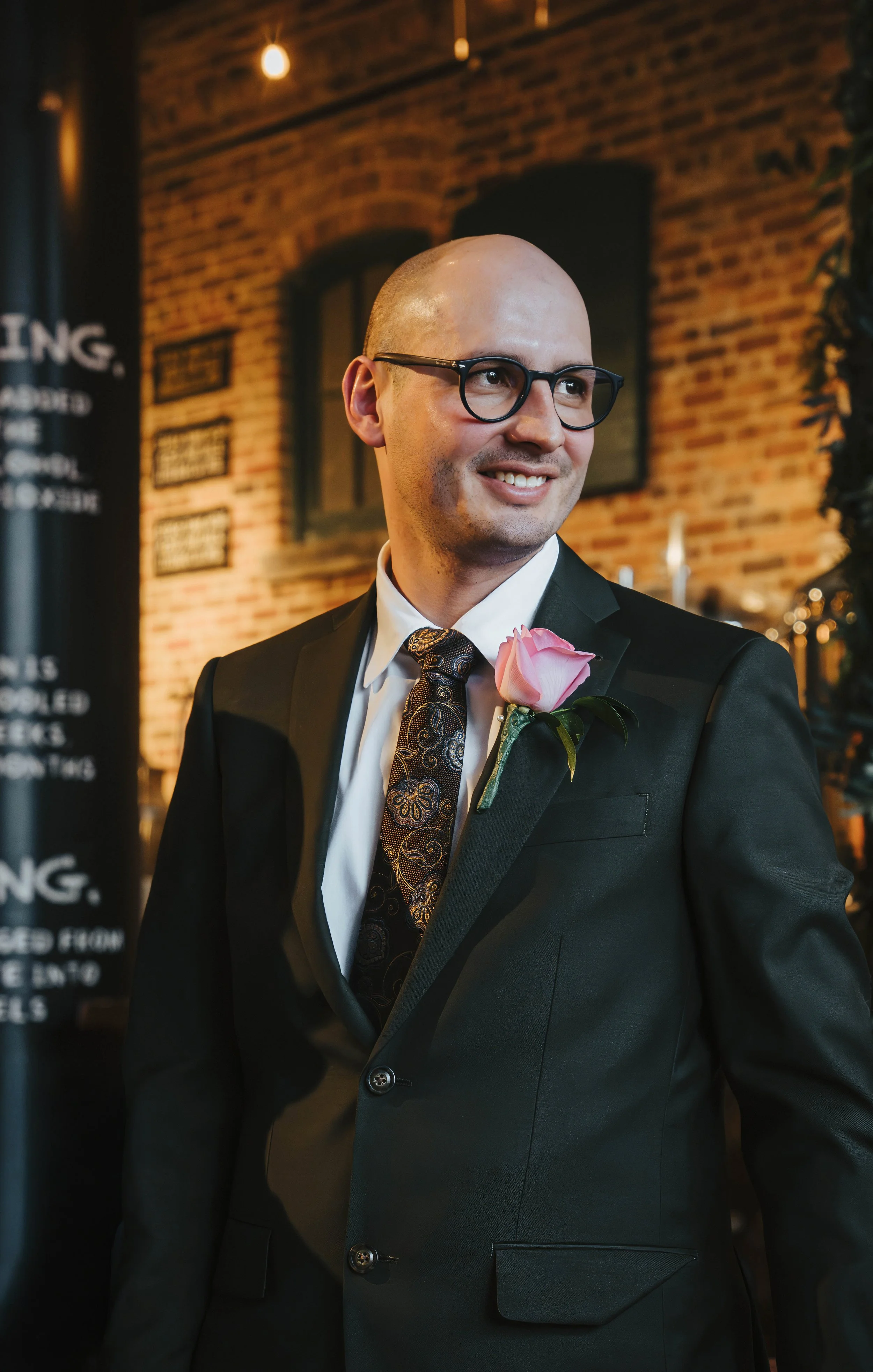 A man in a black suit with a pink flower on his lapel, white shirt, and patterned tie, wearing glasses, smiling in a brick-walled room with warm lighting.