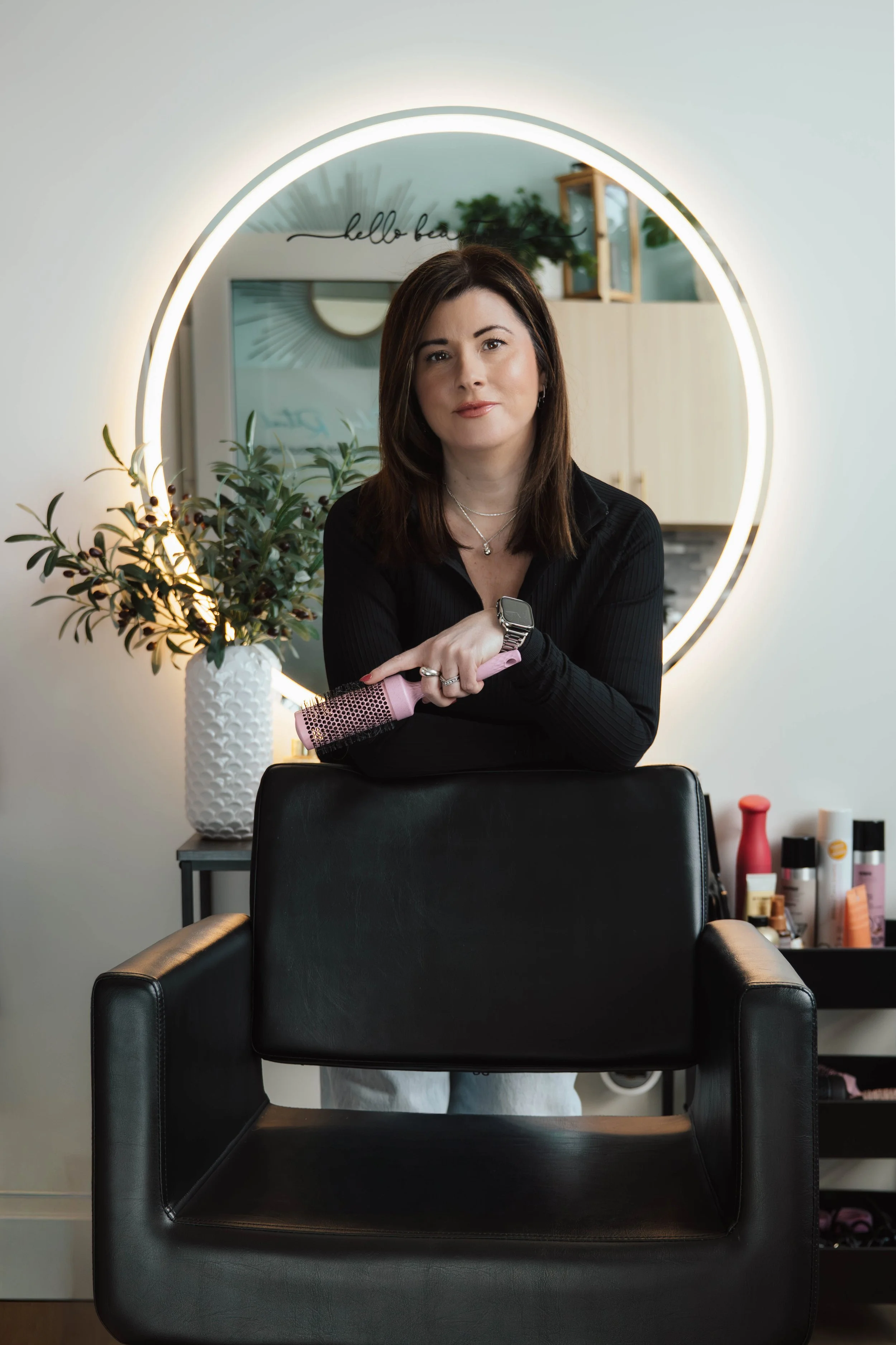 A woman with shoulder-length brown hair stands behind a black salon chair, holding a pink hairbrush, in a salon with a large round mirror with neon lighting, a potted plant, and various hair products in the background.