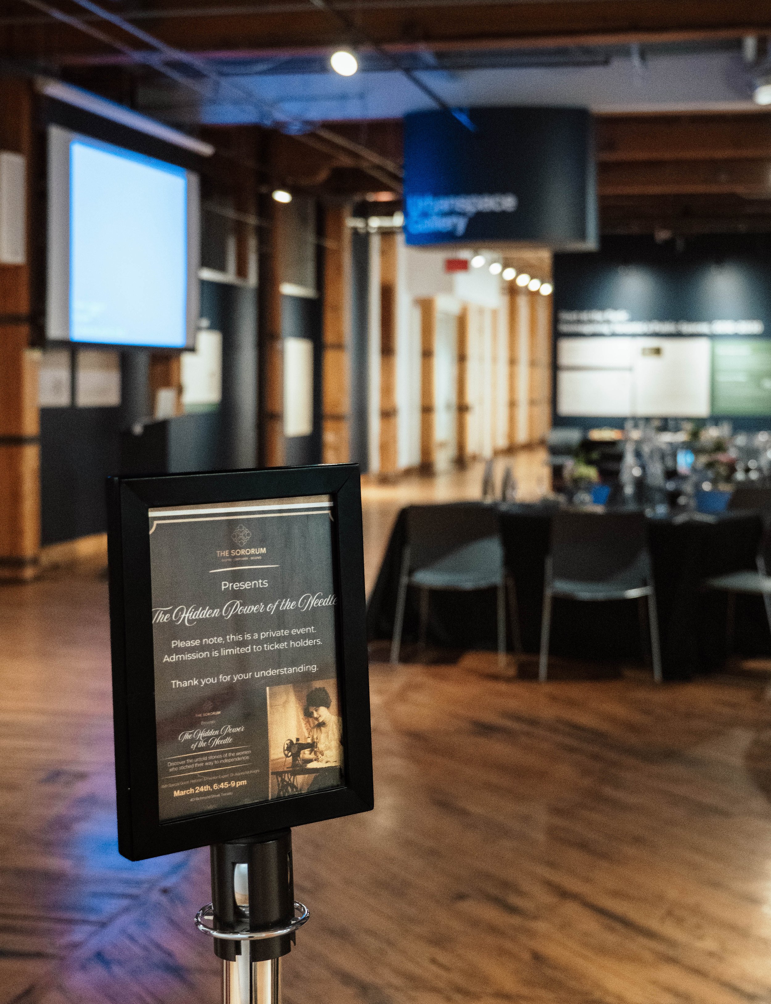 Signboard announcing a private event, 'The Hidden Power of the Needle,' scheduled for March 24th from 6:45 to 9 pm, at The Sororum, with a vintage image of a woman sewing.
