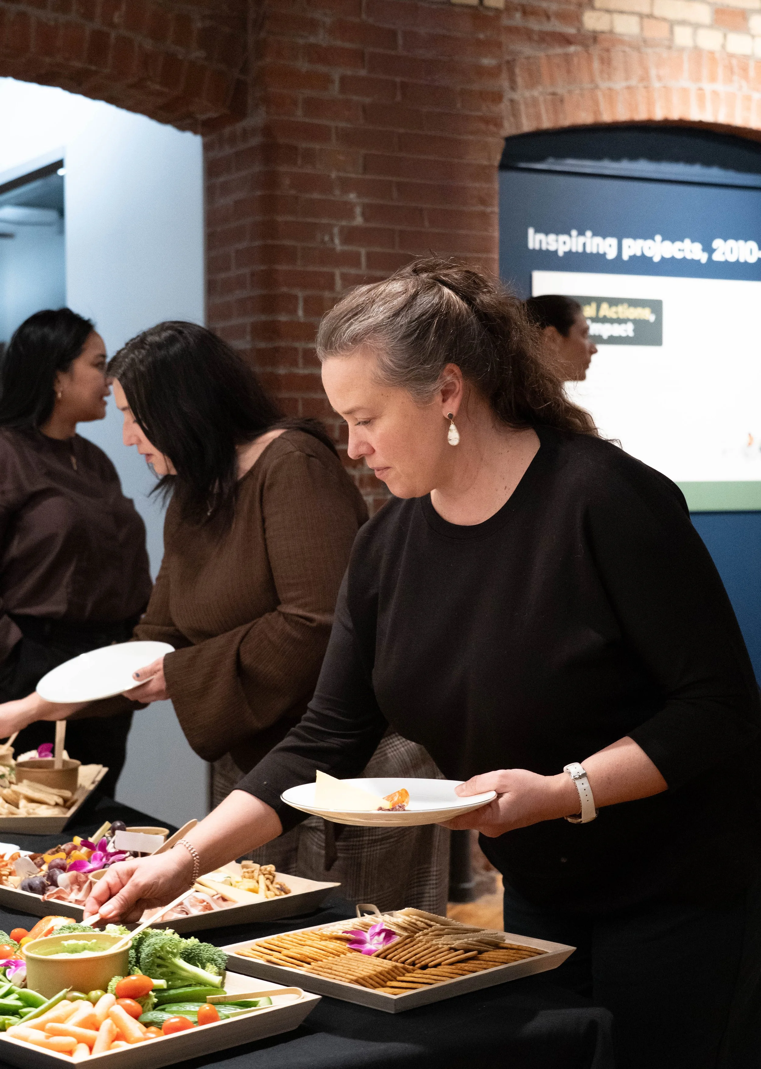 Women serving themselves on a buffet table with vegetables, crackers, and assorted foods at an indoor event.